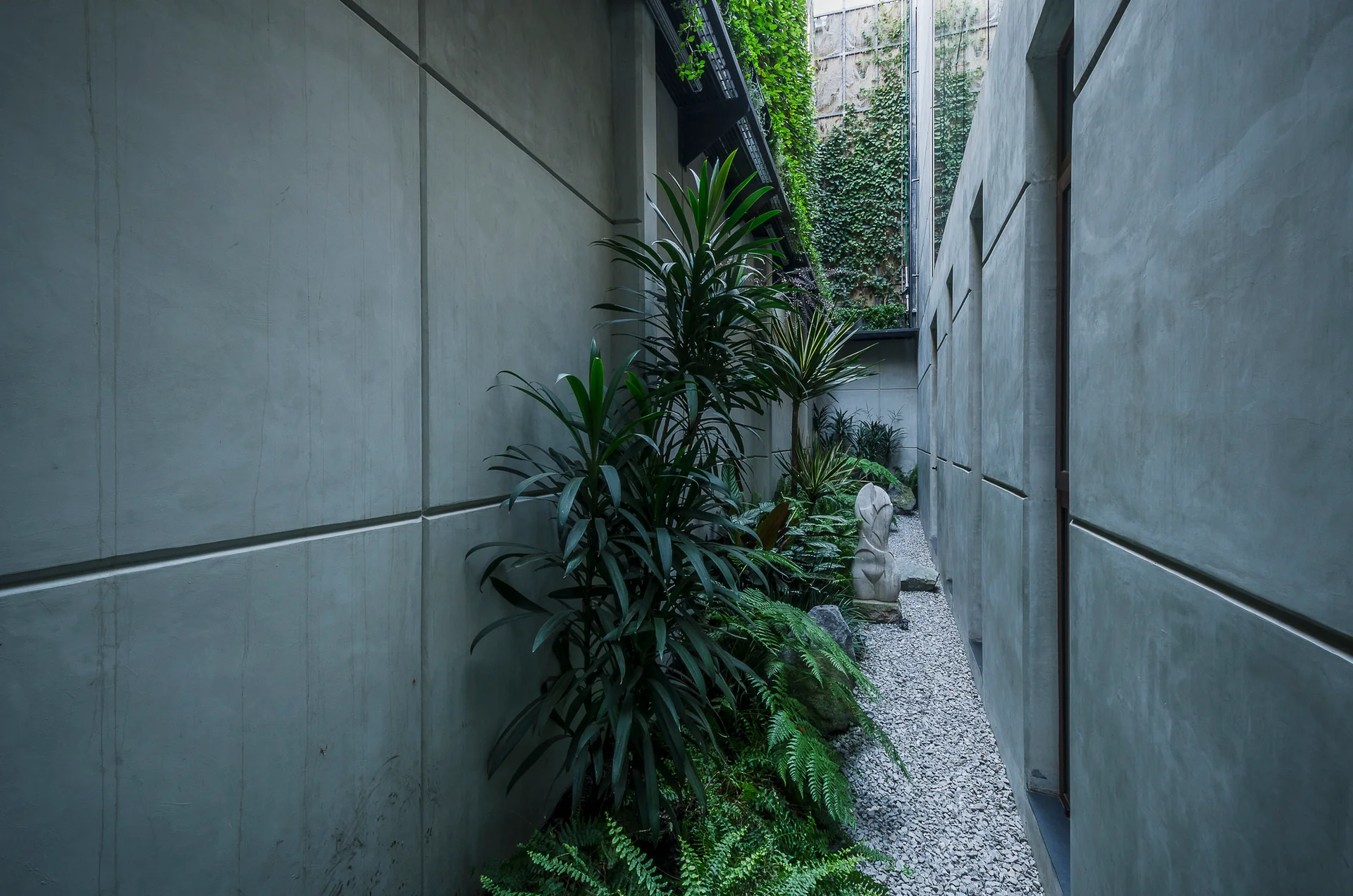 A narrow outdoor corridor features grey concrete paneled walls, a gravel pathway, green ferns, and an abstract stone sculpture.