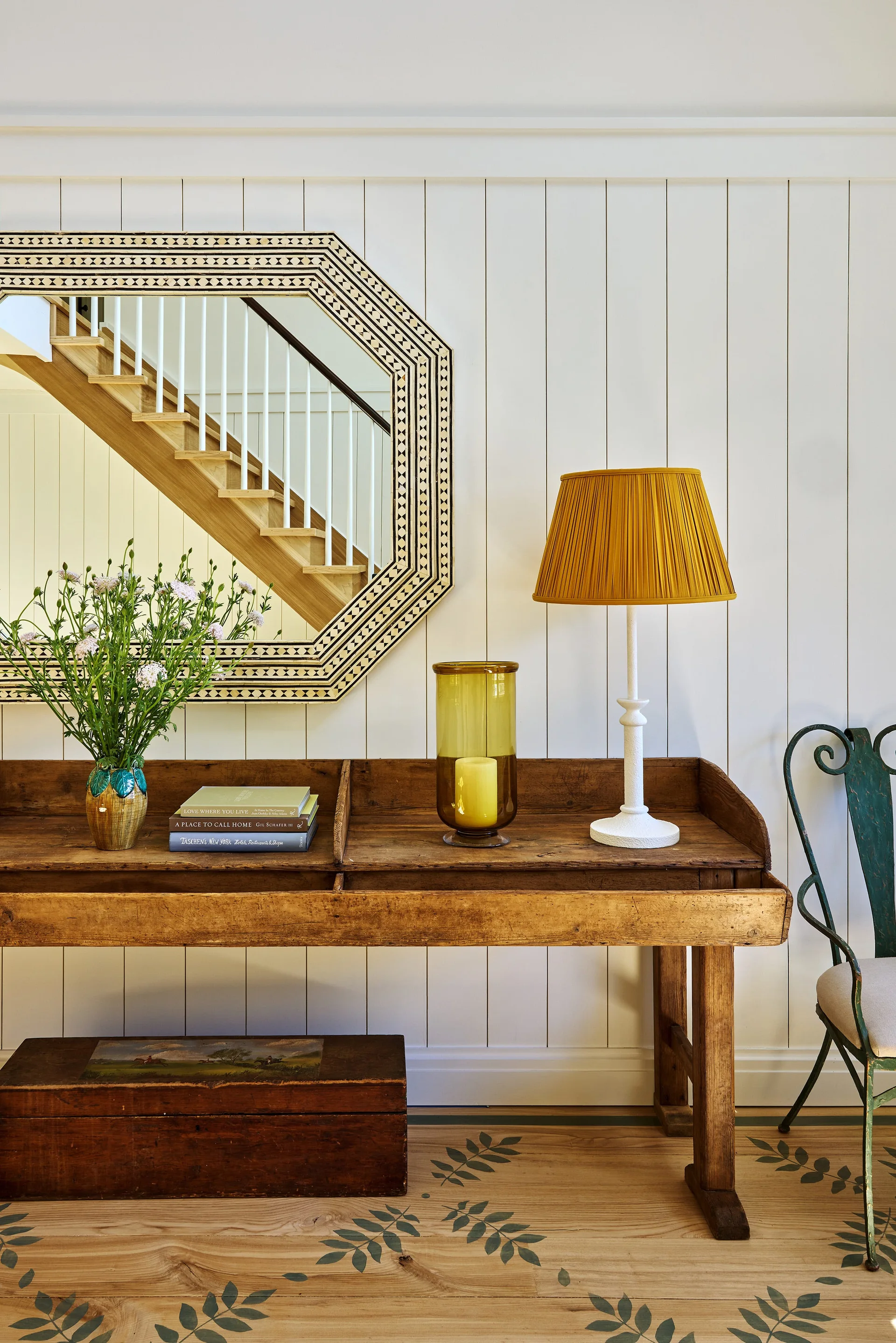 An entryway features a rustic wooden console table beneath an octagonal mirror reflecting stairs, alongside a yellow table lamp and stenciled wood floors.