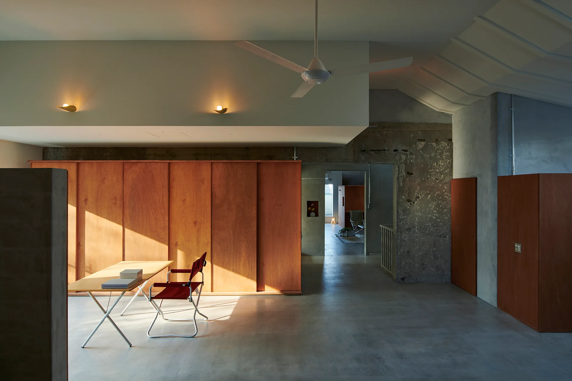 A minimalist home office features raw concrete walls, a large wood-paneled partition, and a simple desk with a red folding chair.