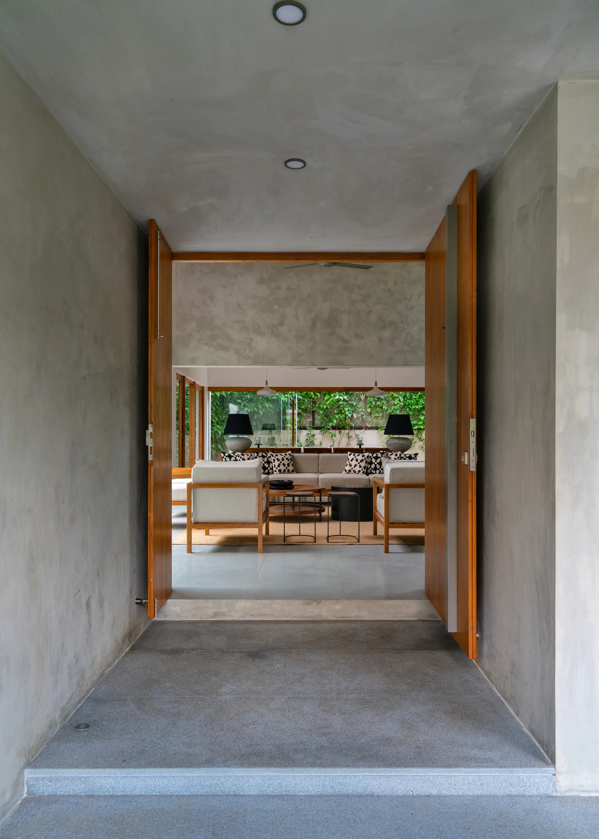 A view through a raw concrete corridor with open wooden pivot doors looking into a modern living room with beige furniture and a green garden view.