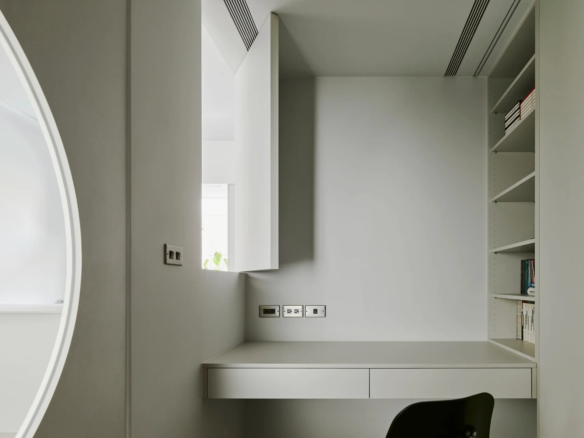 A minimalist study nook features an off-white floating desk, integrated wall shelving, and a circular wall cutout.