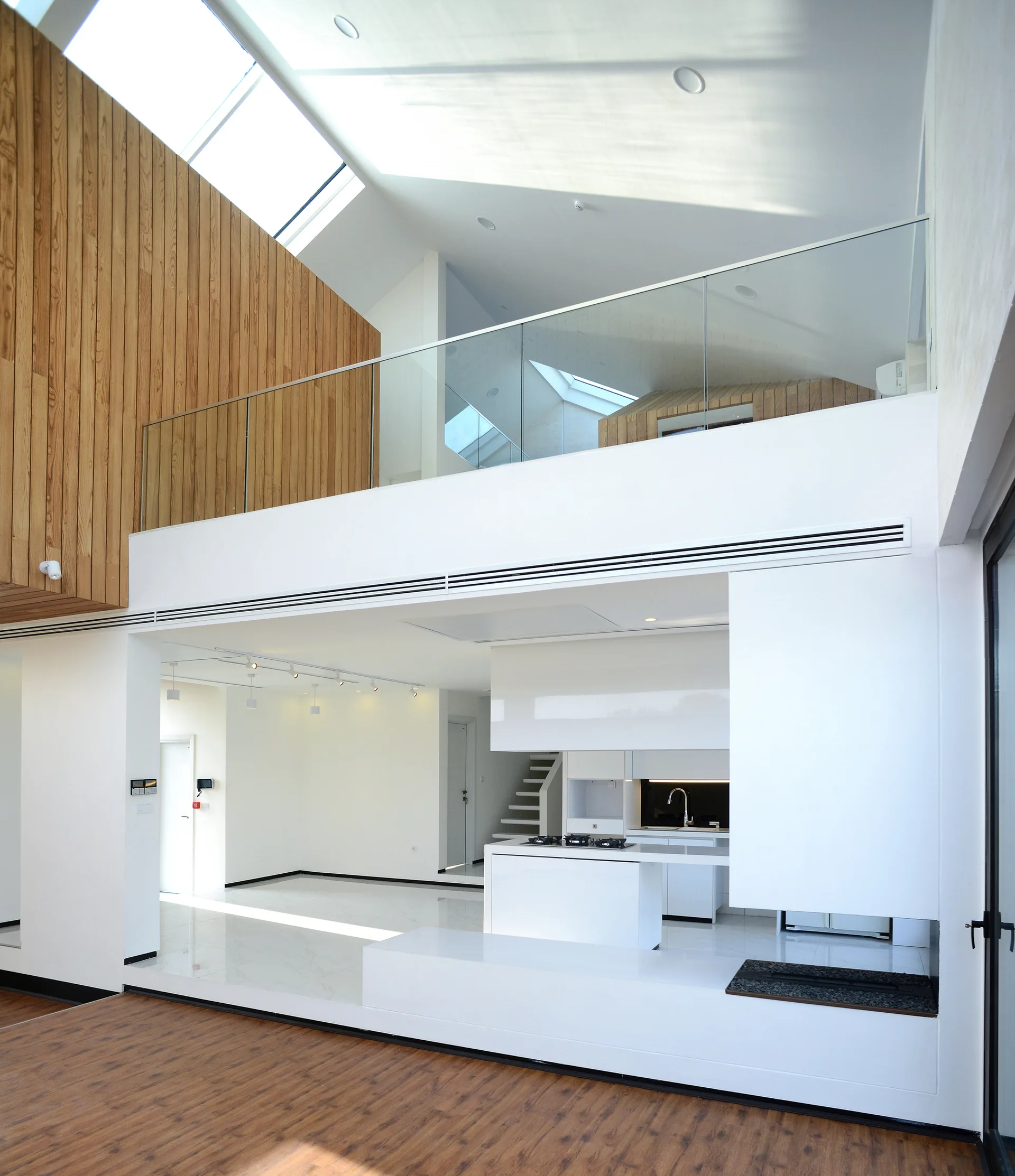 A modern kitchen in a double-height space with white minimalist cabinetry, skylights, a glass mezzanine railing, and vertical wood wall cladding.