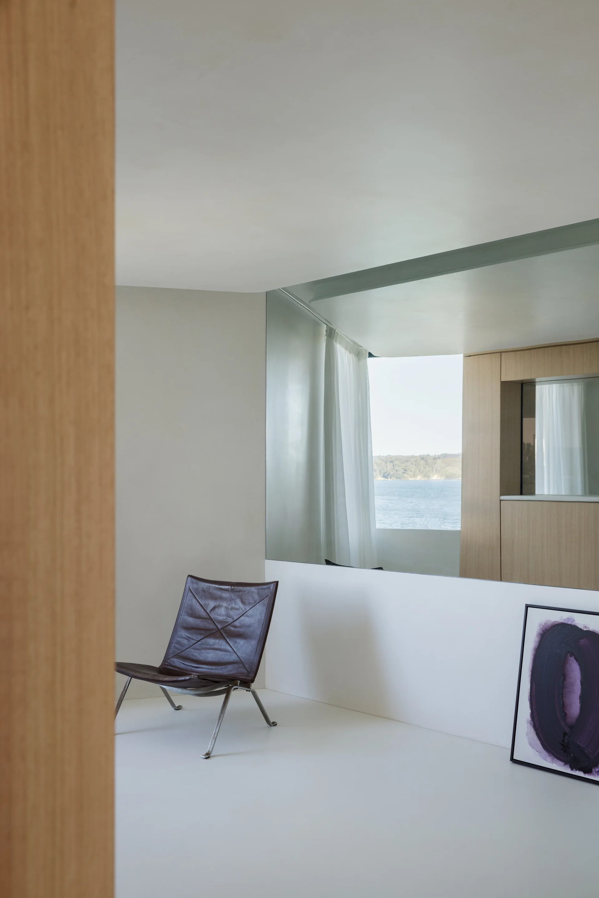 A minimalist room features a brown leather lounge chair on a smooth white floor next to a large mirror reflecting a sea view window.