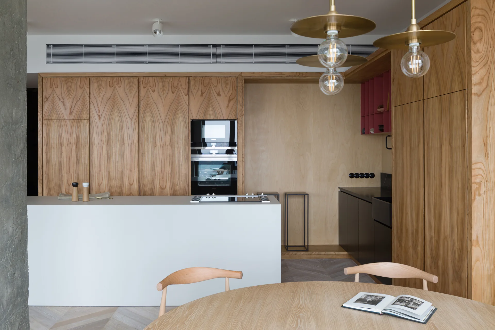 A modern kitchen features light wood cabinets, a white minimalist island with a built-in cooktop, and a round wood dining table.
