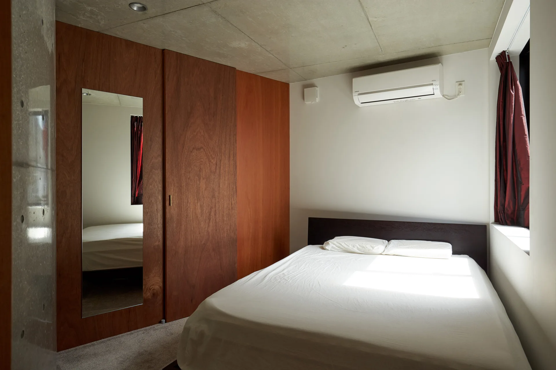 A minimalist bedroom with an exposed concrete ceiling, a white bed, and warm wood-paneled closet doors.