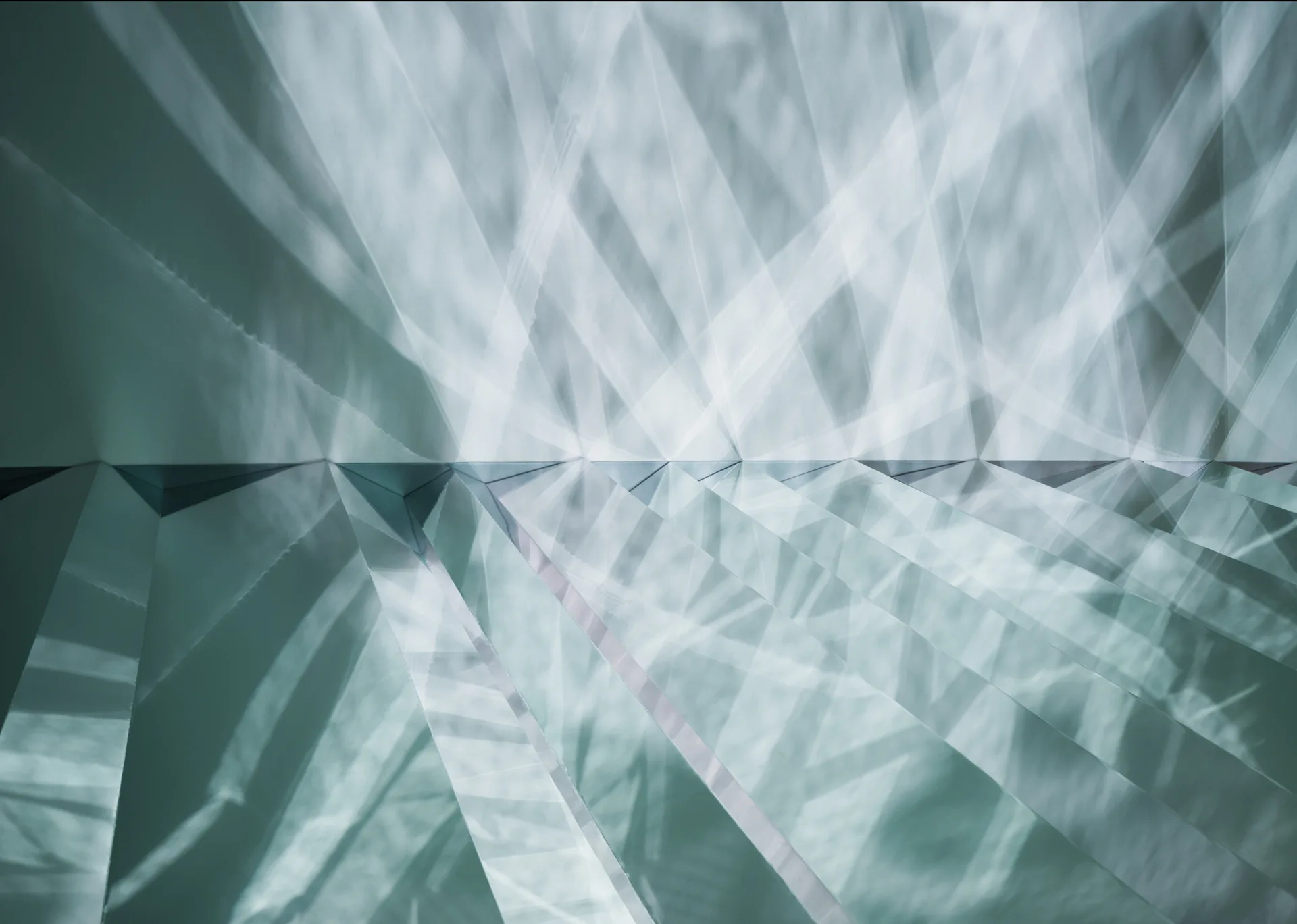 Light reflects across angled, faceted metallic panels on a wall to create sharp geometric streaks of white and blue-grey shadows.