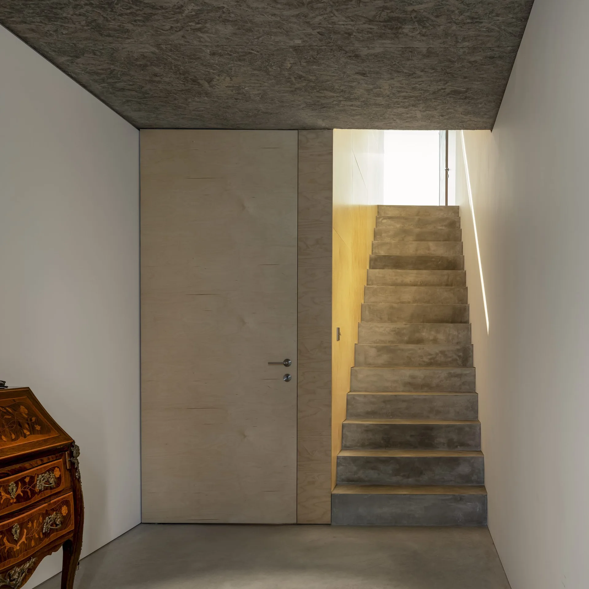 A minimalist entryway features cast concrete stairs ascending next to a flush plywood door and wall underneath a dark textured ceiling.