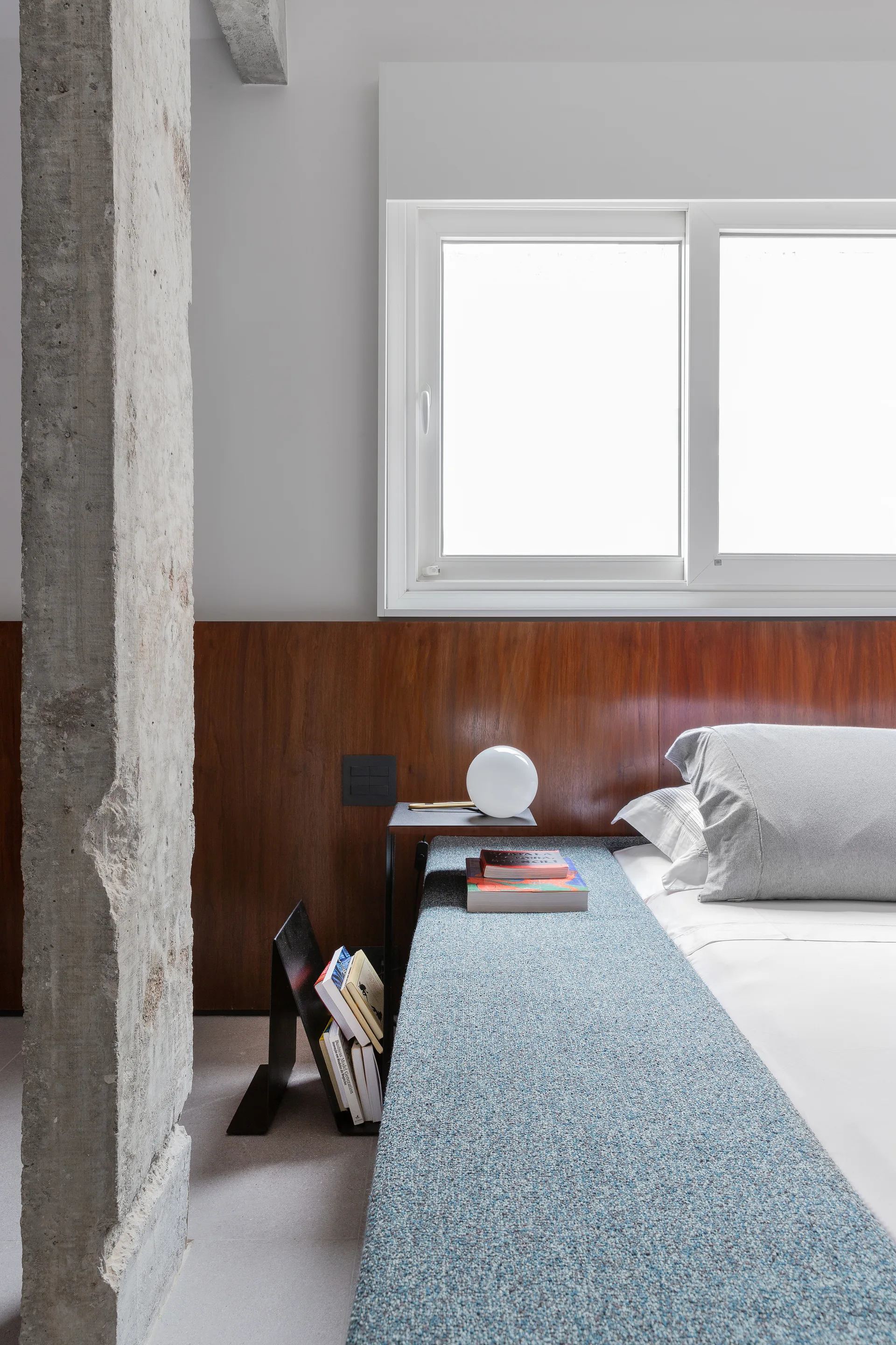A modern bedroom with grey bedding, a warm wood wall panel headboard, an exposed concrete column, and a small bedside table with a globe lamp.