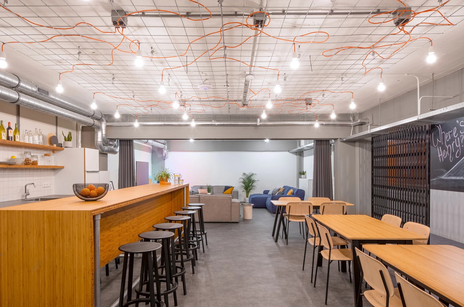 An industrial dining space with a wooden bar, dining tables, grey concrete floors, and a ceiling grid woven with orange electrical cords.
