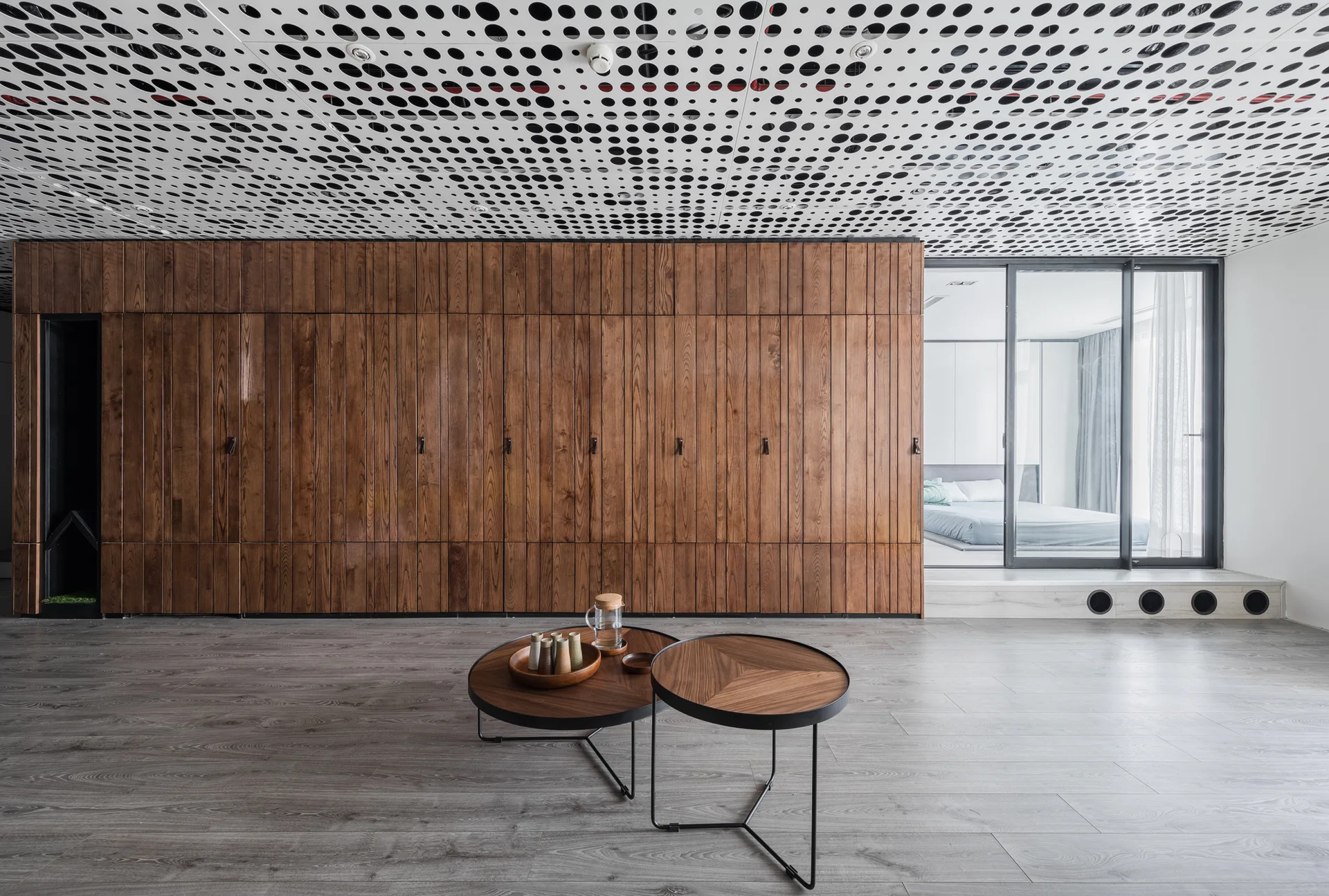 A minimalist living room features nested wooden coffee tables, a large wood-paneled storage wall, and a white perforated ceiling.