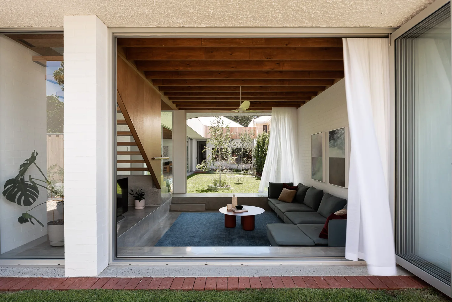 A sunken living room with a dark grey sectional sofa, white brick walls, and exposed wood ceiling beams opens to a grassy courtyard.