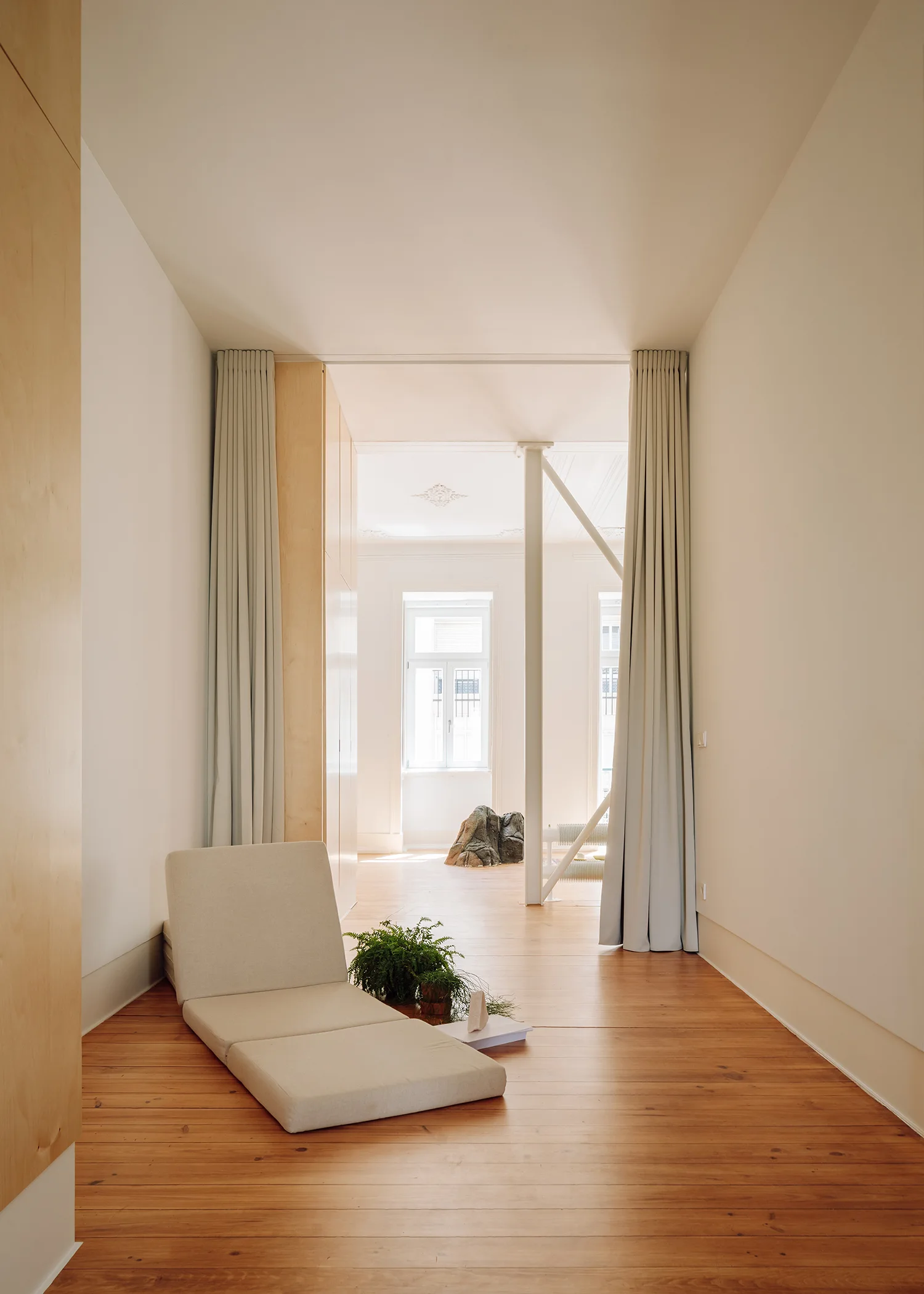 A minimalist interior features a low beige floor chair, a potted fern on wide-plank wood floors, and floor-to-ceiling curtains framing a doorway.