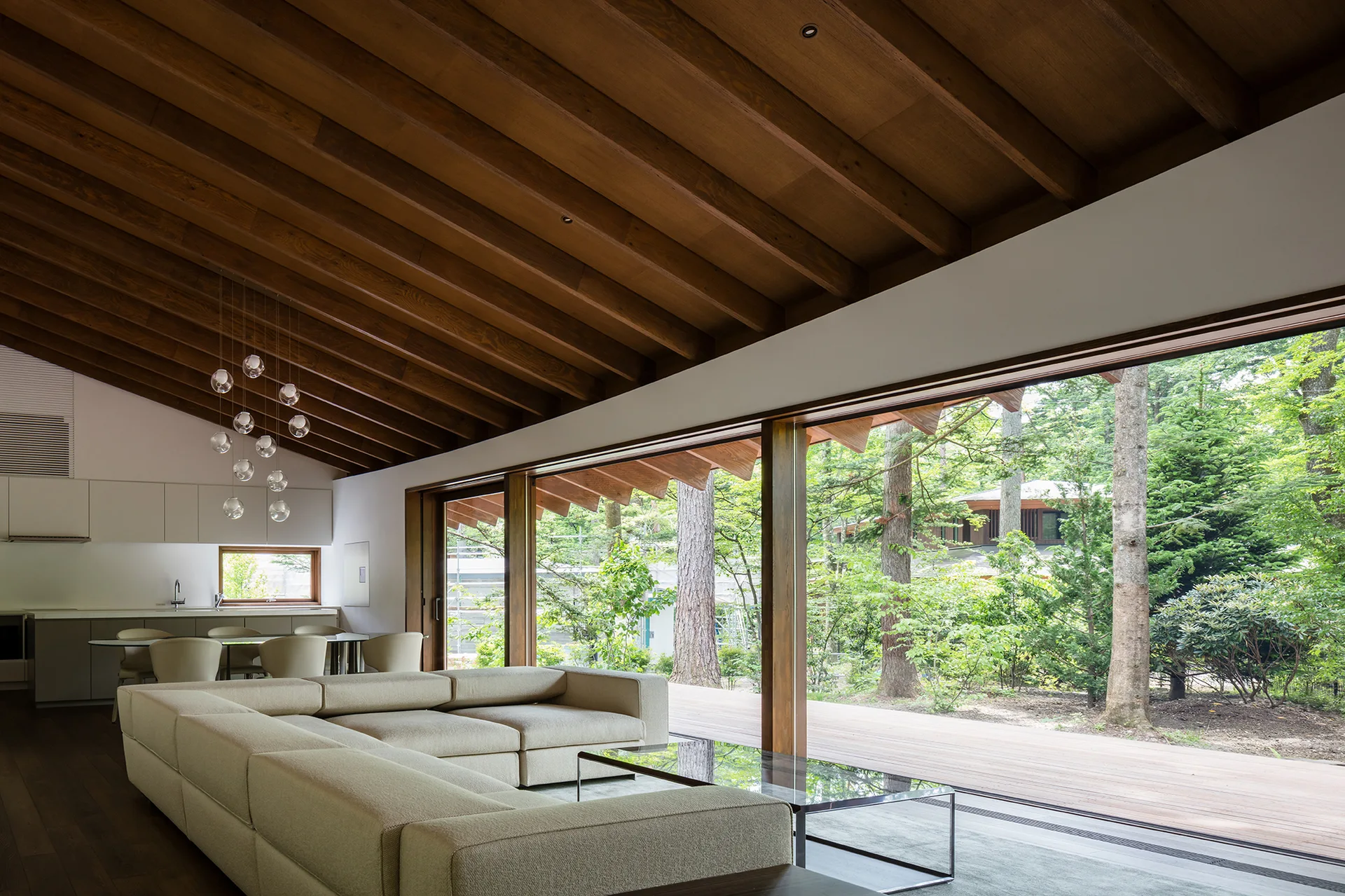 A modern living room features a cream sectional sofa, a glass coffee table, and an exposed wood ceiling beside sliding glass doors opening to a forest.