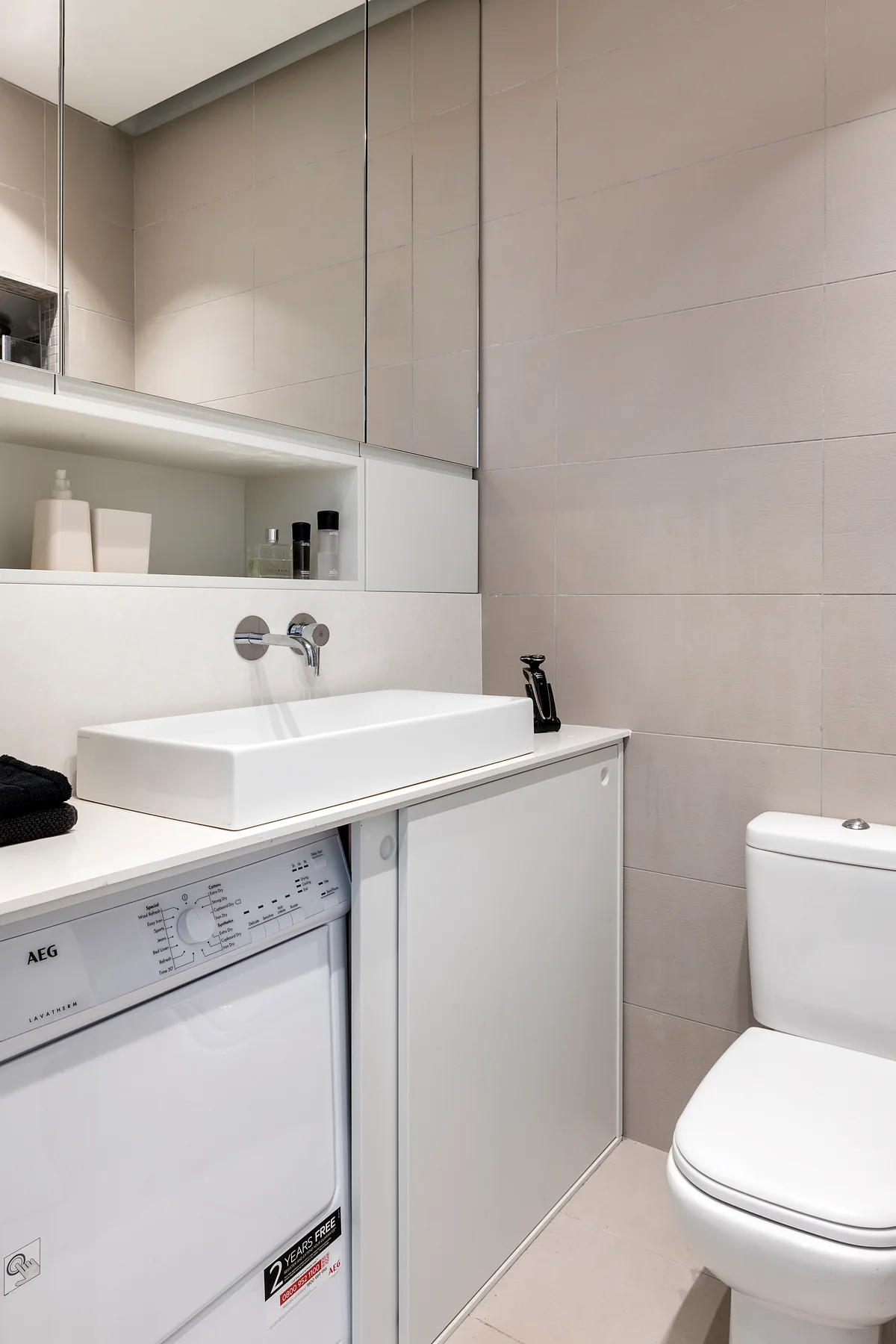 A compact modern bathroom features a white vanity unit housing a washing machine beneath a wall-mounted sink and a mirrored cabinet.