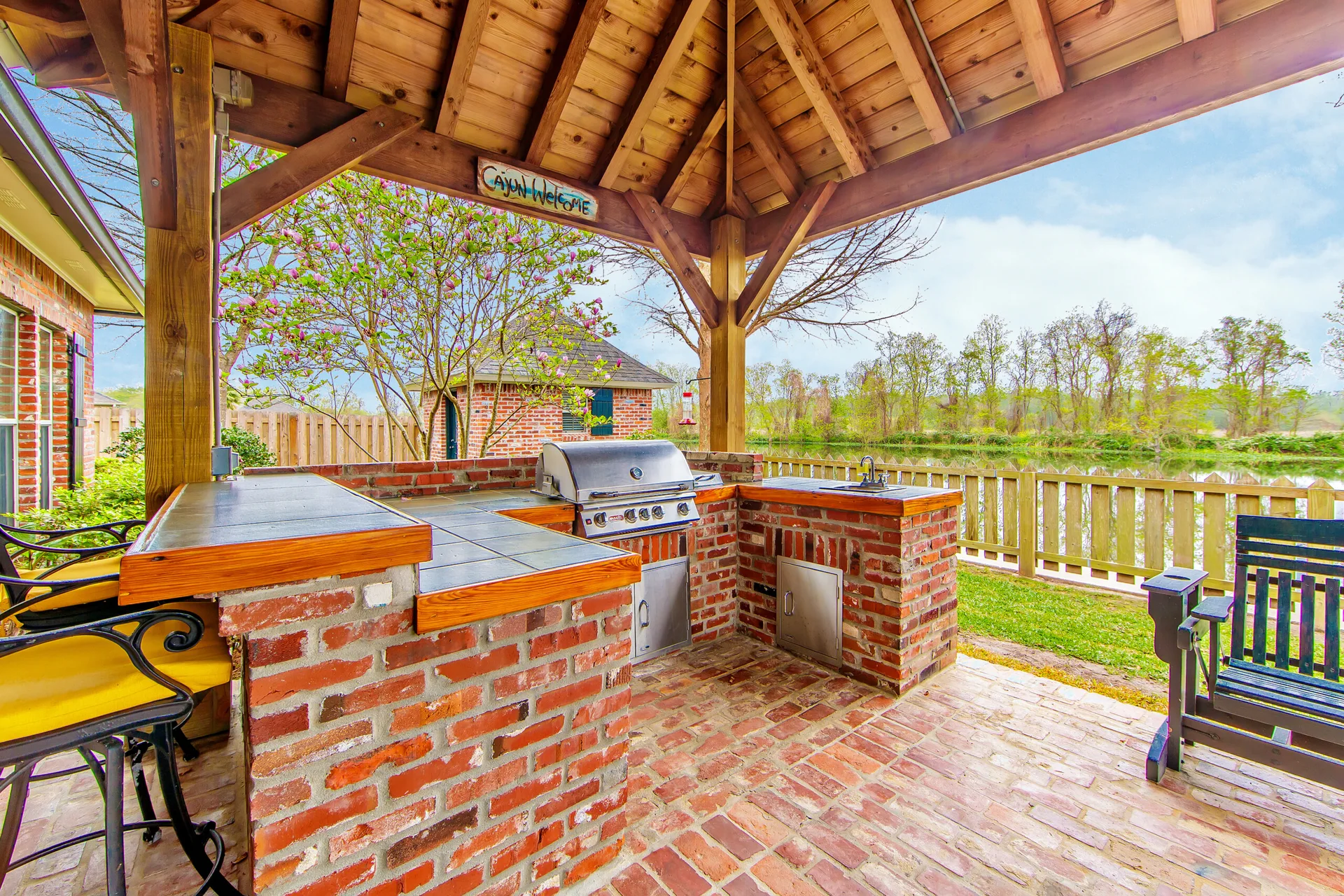 A rustic outdoor kitchen built with red brick features a stainless steel grill and sink under a high timber pavilion ceiling next to a lake.