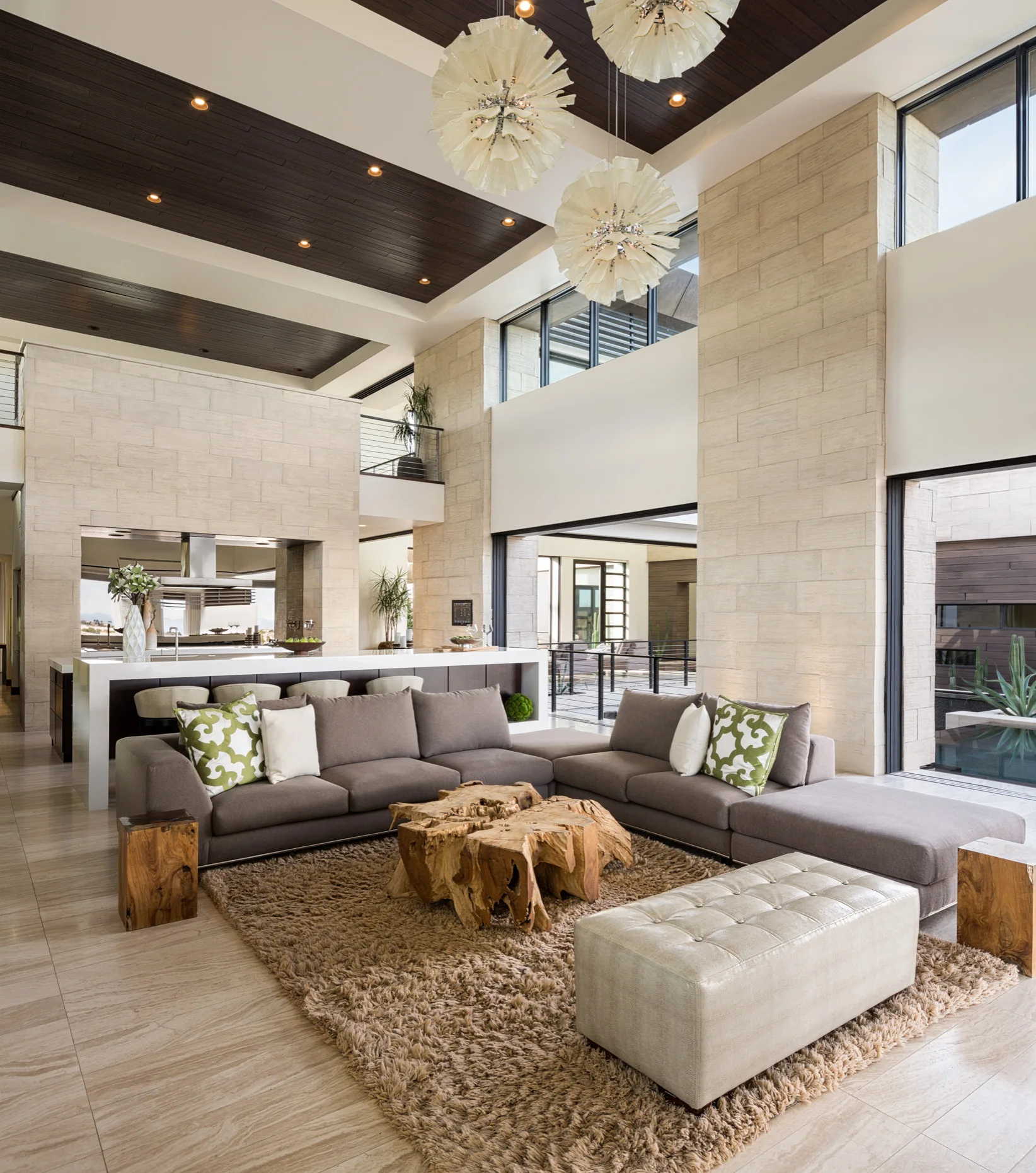 A spacious living room features a grey sectional sofa, an organic root coffee table, a shag rug, and tall stone tile walls under a dark wood ceiling.