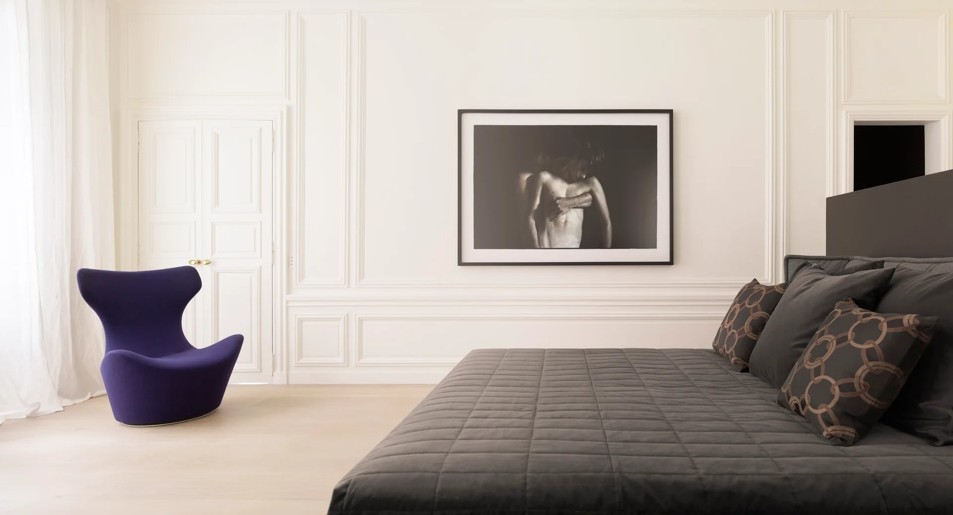 A minimalist bedroom featuring white paneled walls, a large grey quilted bed, a deep purple organic accent chair, and framed black-and-white art.