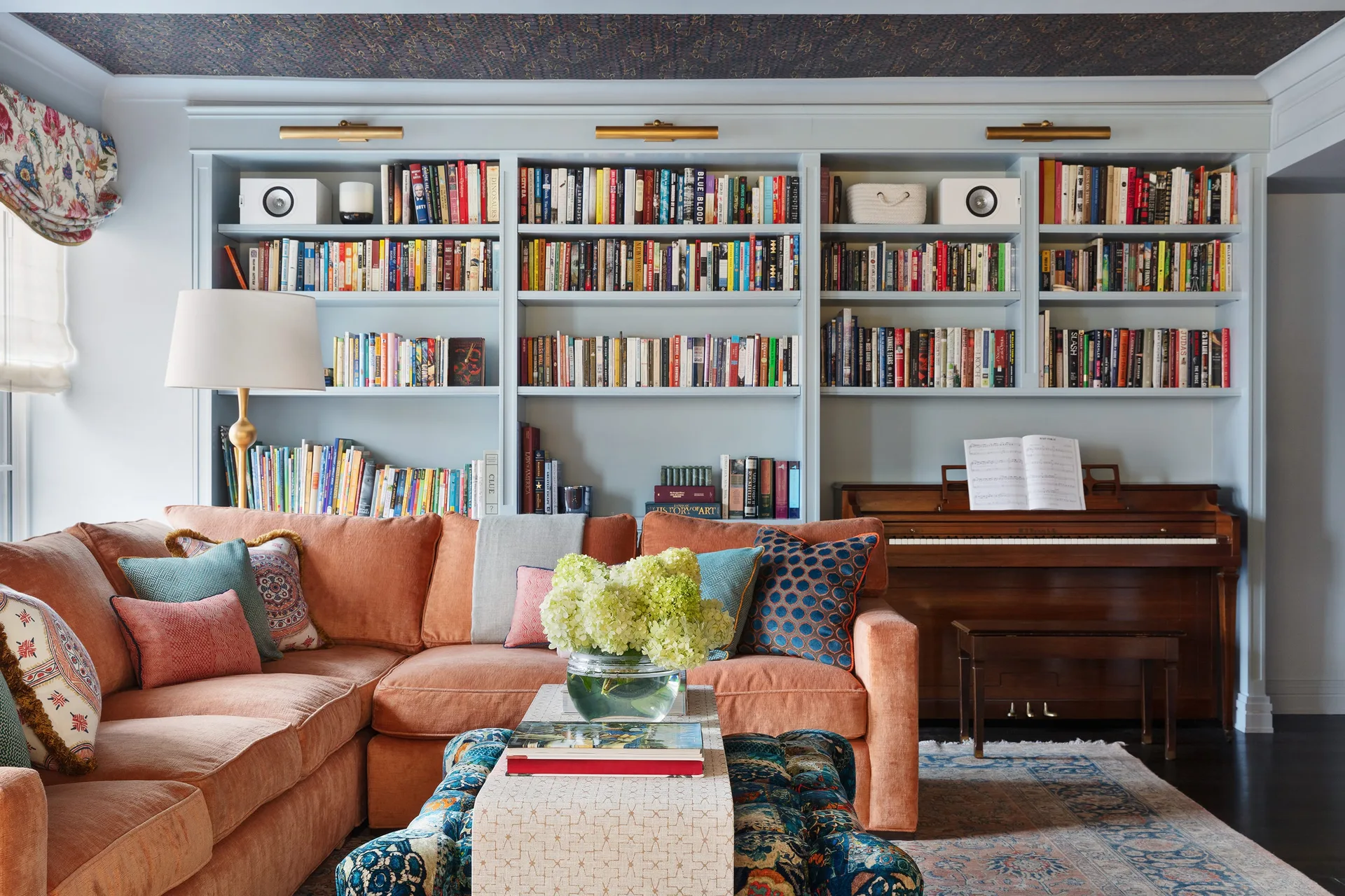 A cozy living room featuring a large light blue built-in bookshelf wall, an integrated upright wood piano, and an orange sectional sofa.