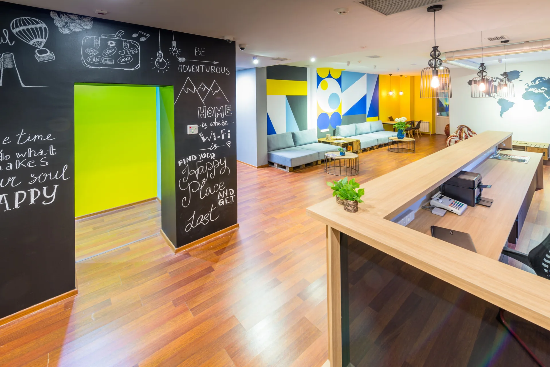 A modern coworking reception space features a wood-toned desk, a chalkboard wall with hand-drawn art, and grey lounge sofas in the background.