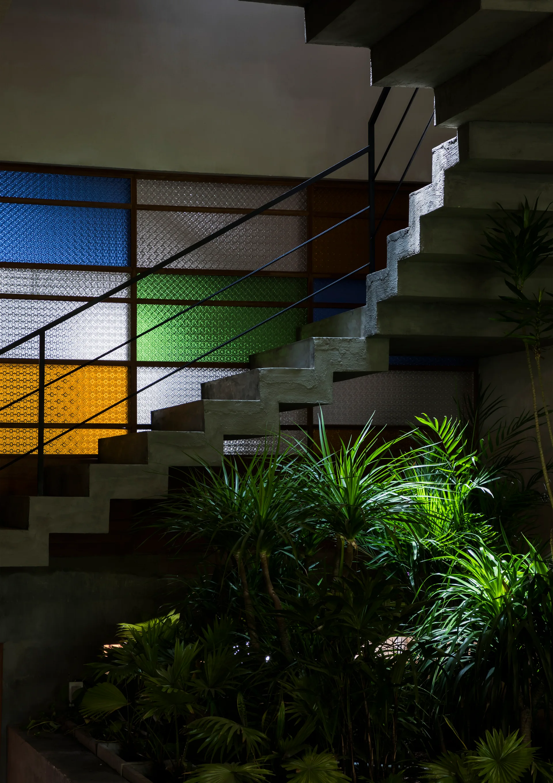 A concrete floating staircase ascends past a geometric grid of yellow, green, and blue stained glass windows with leafy green plants growing below.