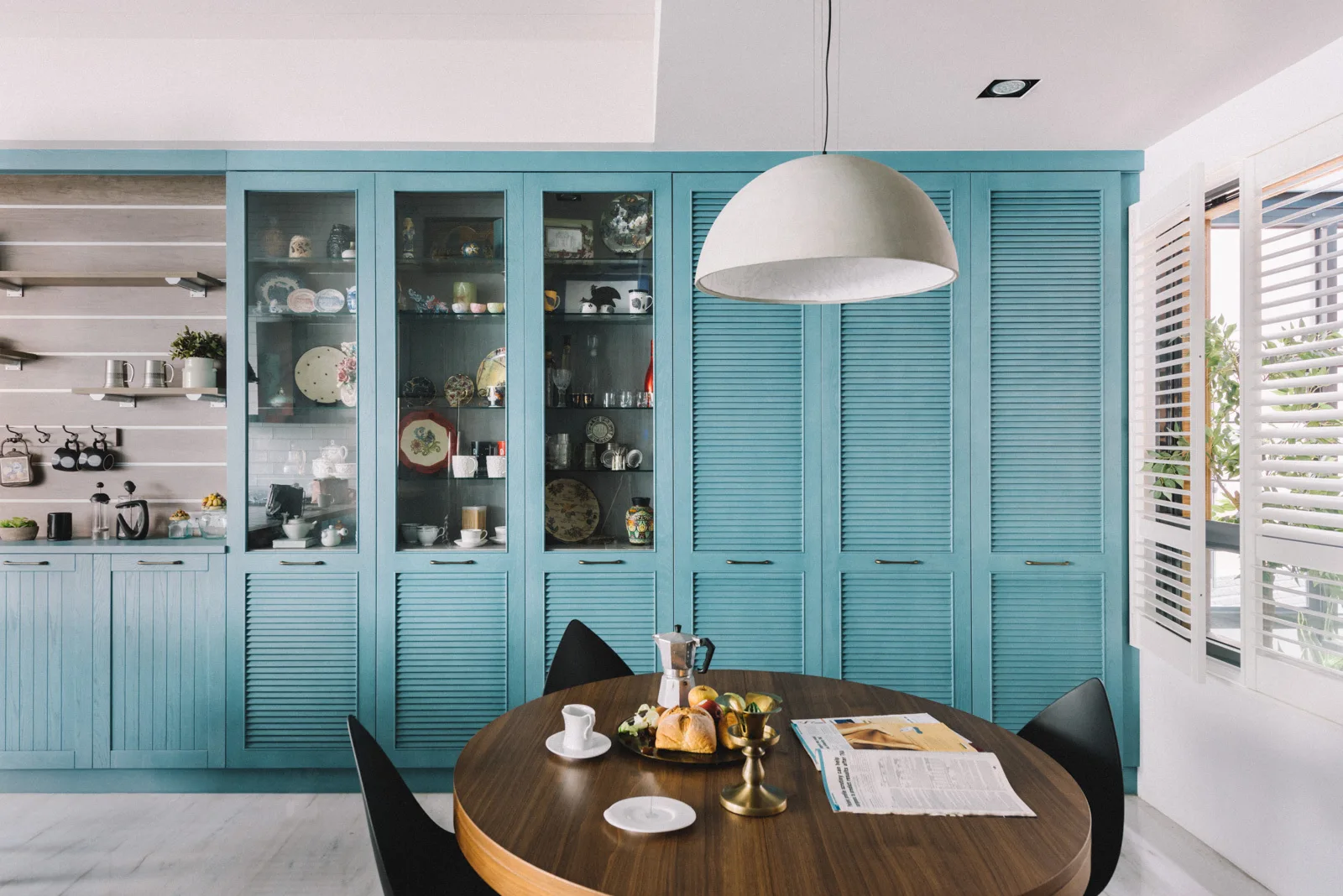 A round wooden dining table and black chairs stand before a massive wall of teal louvered cabinets and glass-front display cases.