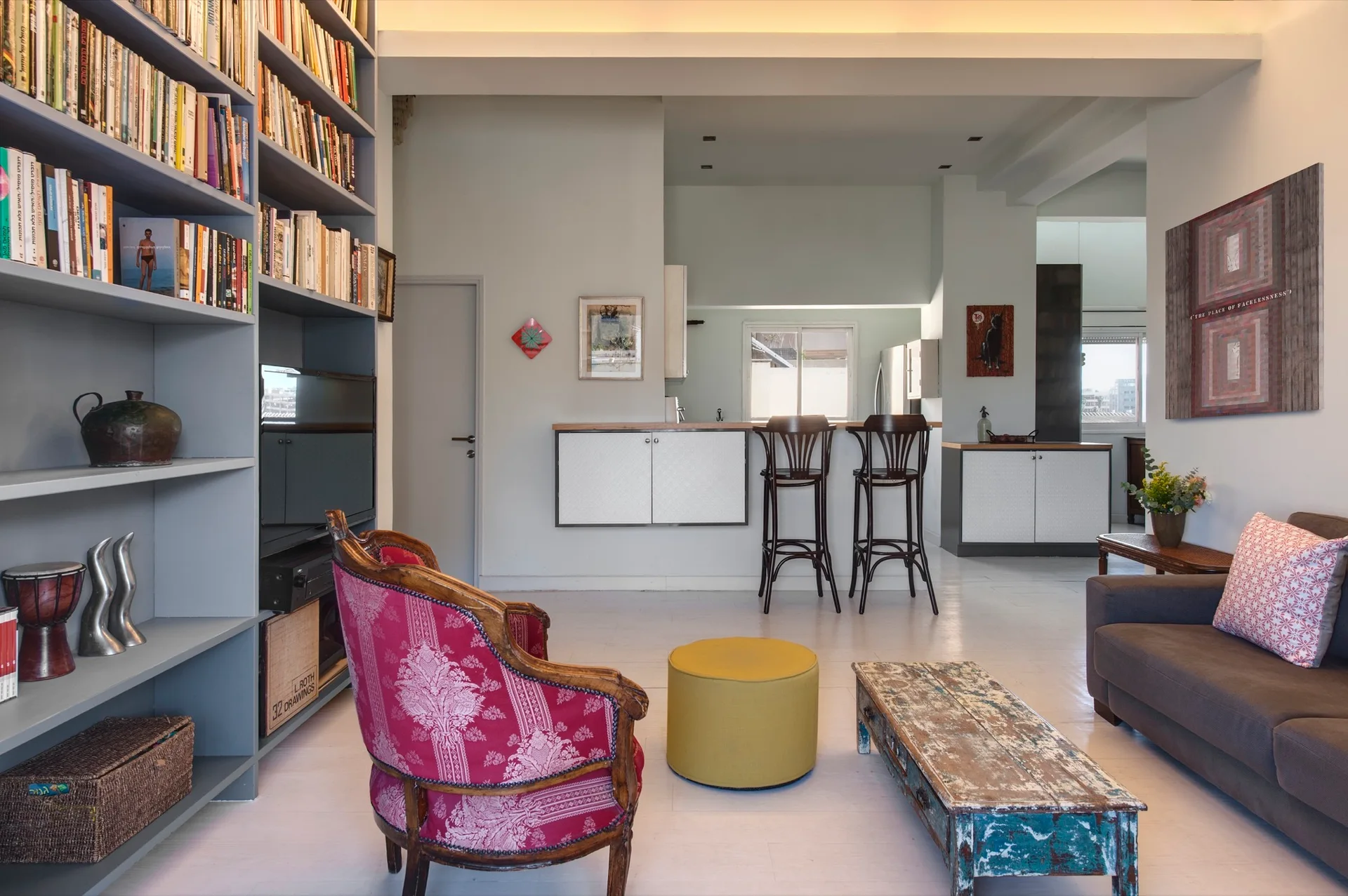 An open concept living room features a large grey bookshelf, a dark grey sofa, a pink patterned armchair, and a distressed wood coffee table.