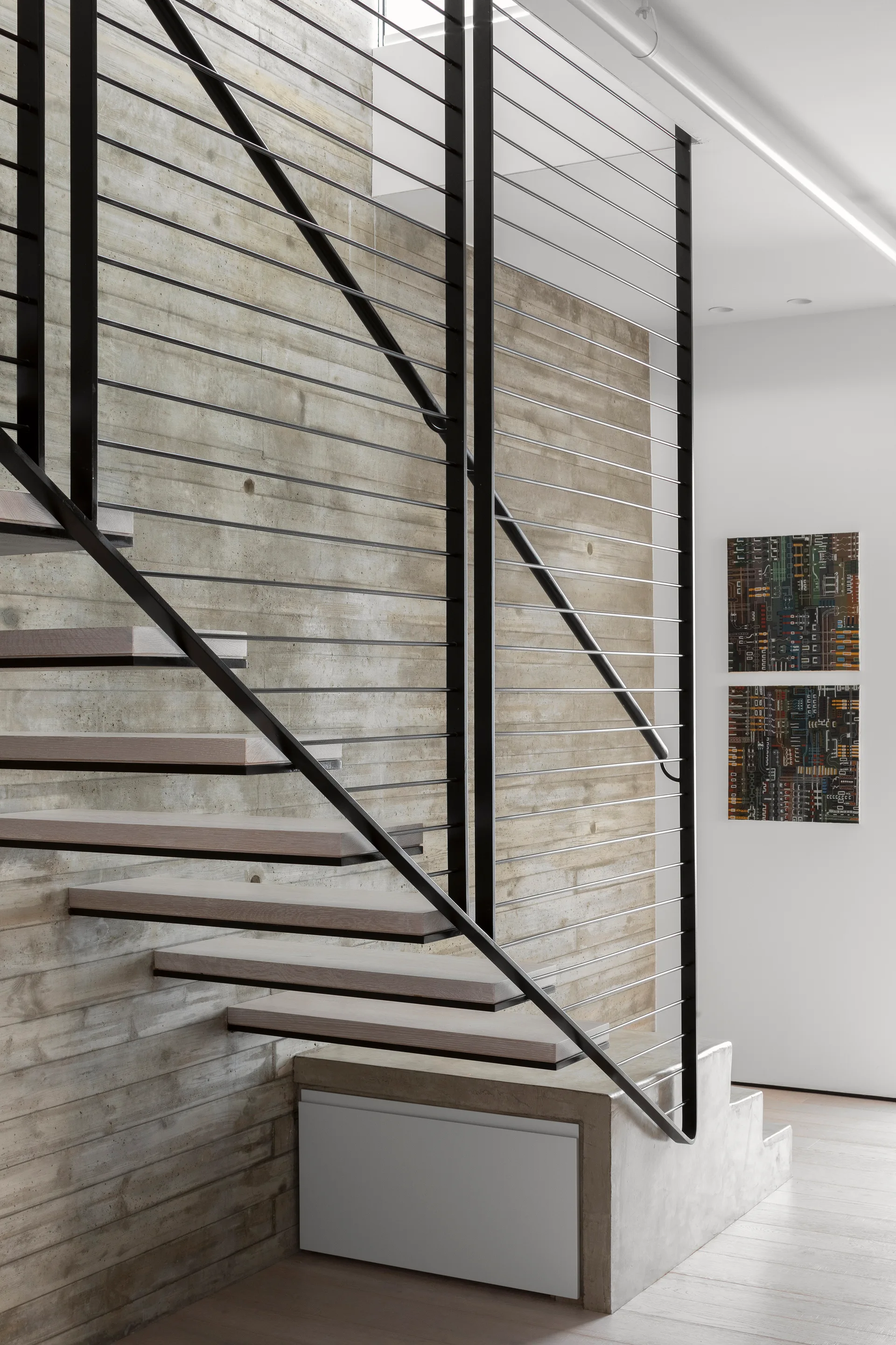 An open riser staircase with light wood treads and a black metal cable railing stands in front of a textured board-formed concrete wall.