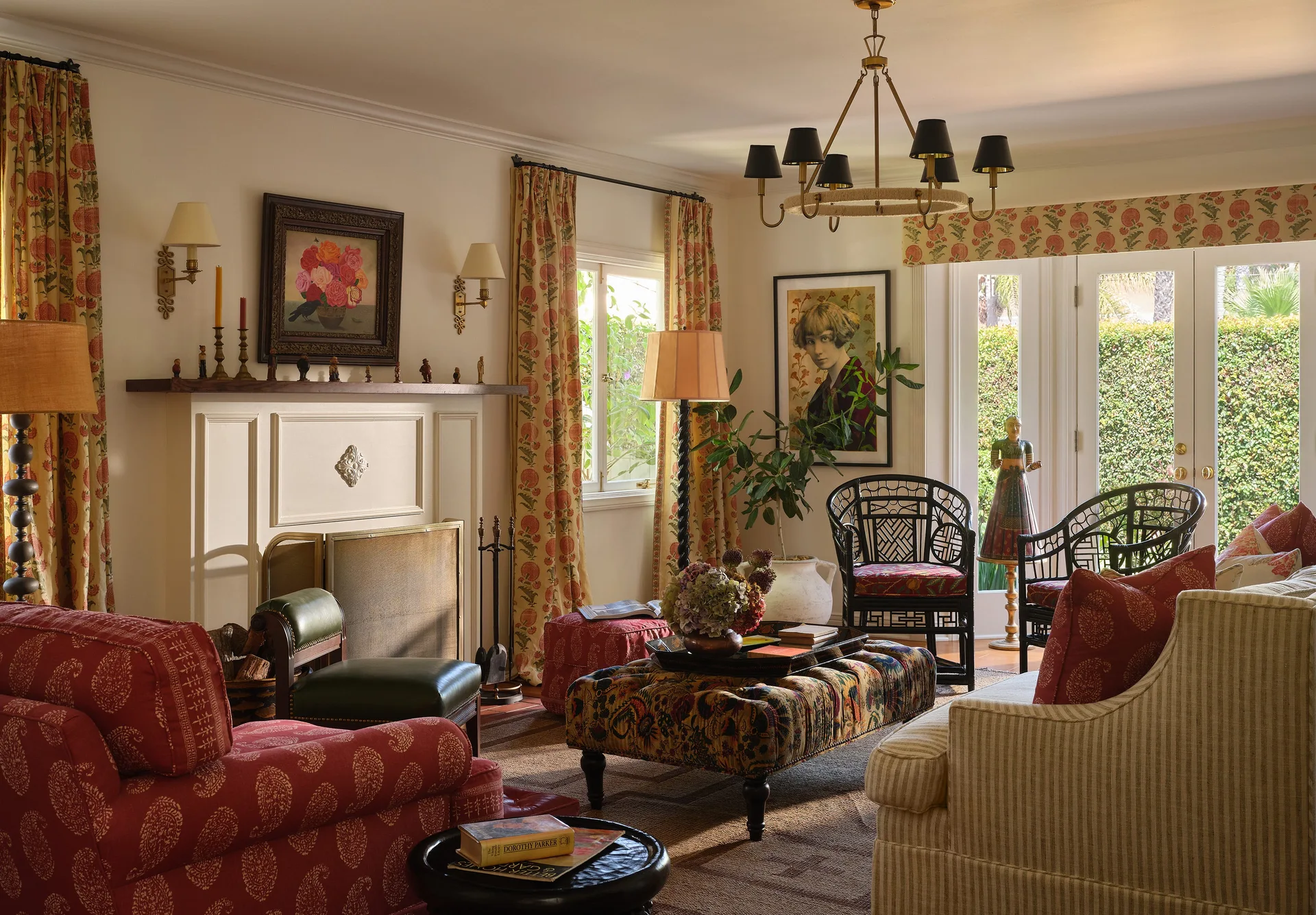 A traditional living room features a fireplace, a red patterned sofa, a floral upholstered ottoman, and black rattan armchairs near glass French doors.
