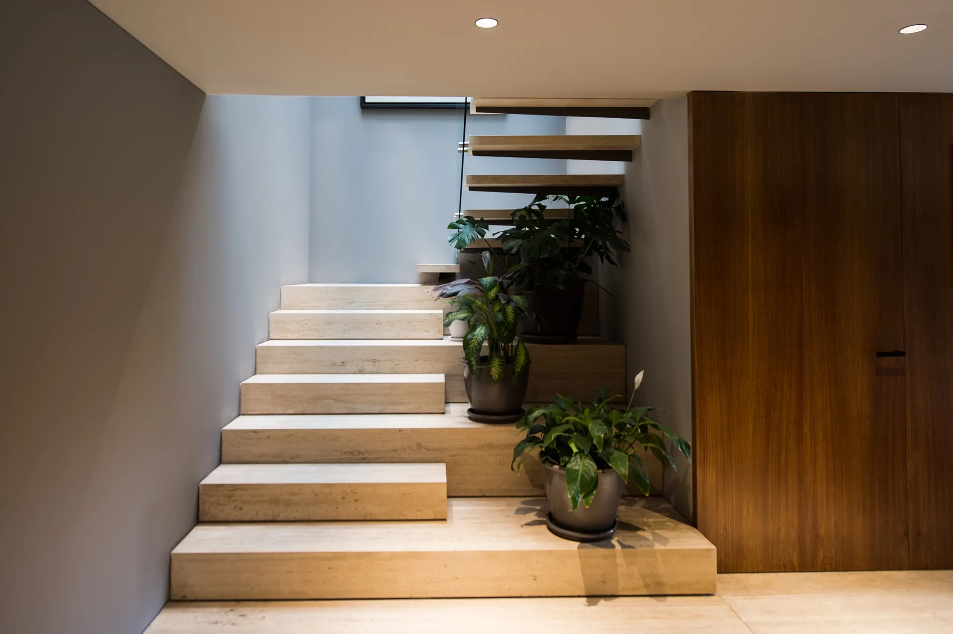 A minimalist beige stone staircase features wide steps styled with potted green plants next to a vertical wood panel wall.