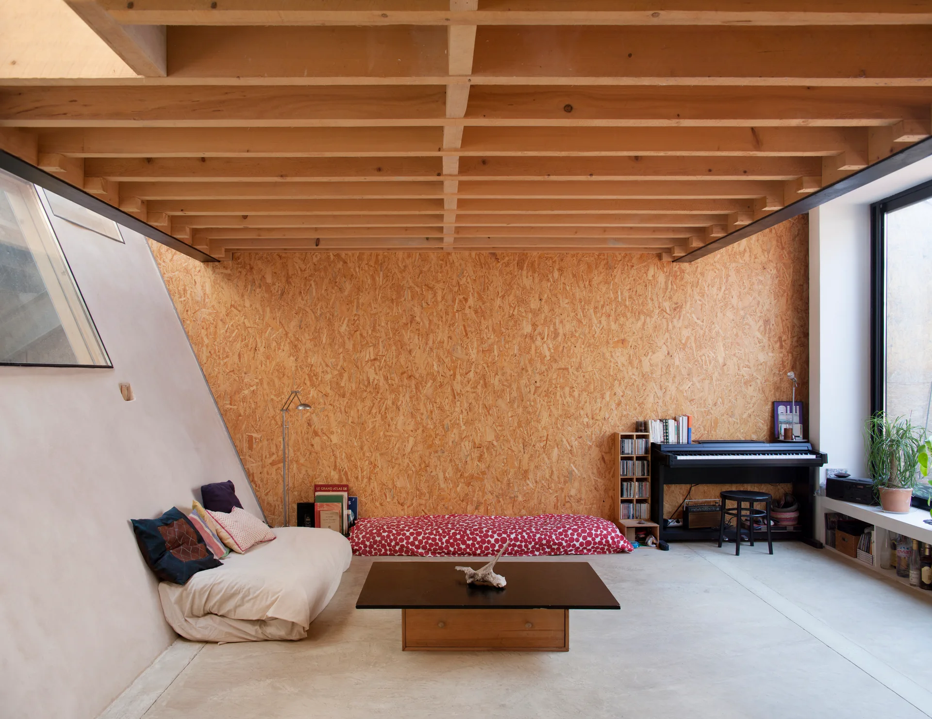 A minimalist loft living room features an OSB accent wall, exposed wooden ceiling joists, concrete flooring, and low-profile seating.
