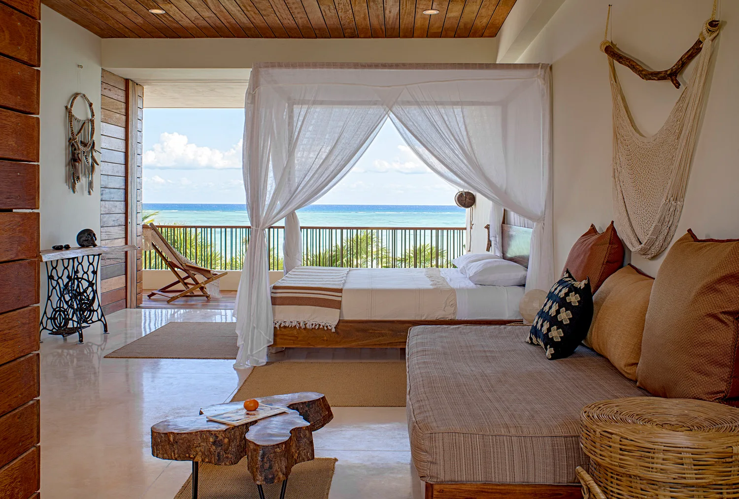 A coastal bedroom features a wooden canopy bed with sheer white drapes, a low sofa, and an open balcony overlooking the ocean.