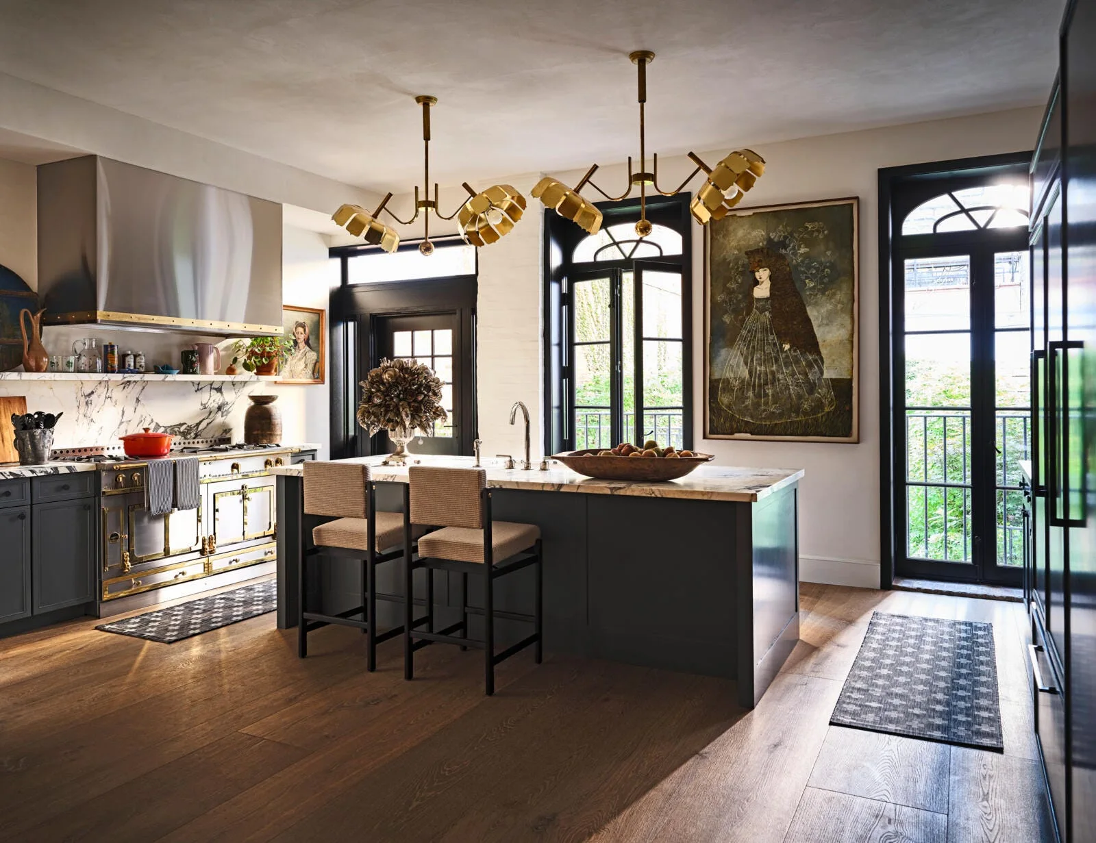 A spacious kitchen features a charcoal island with bar stools, sculptural brass chandeliers, veined marble backsplash, and black arched French doors.