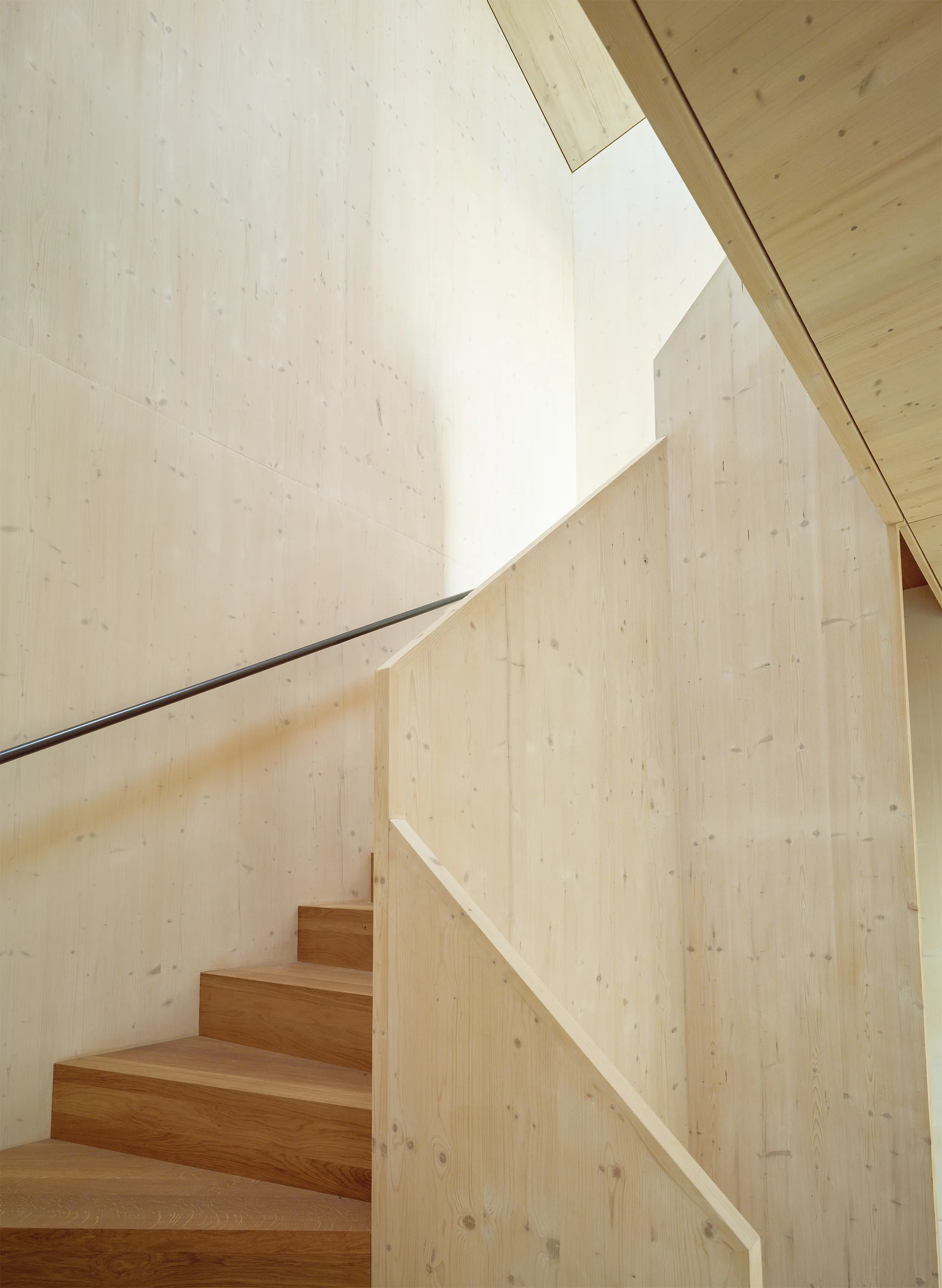 A minimalist wooden staircase features light-colored wall paneling, darker wood treads, and a slim dark metal handrail.