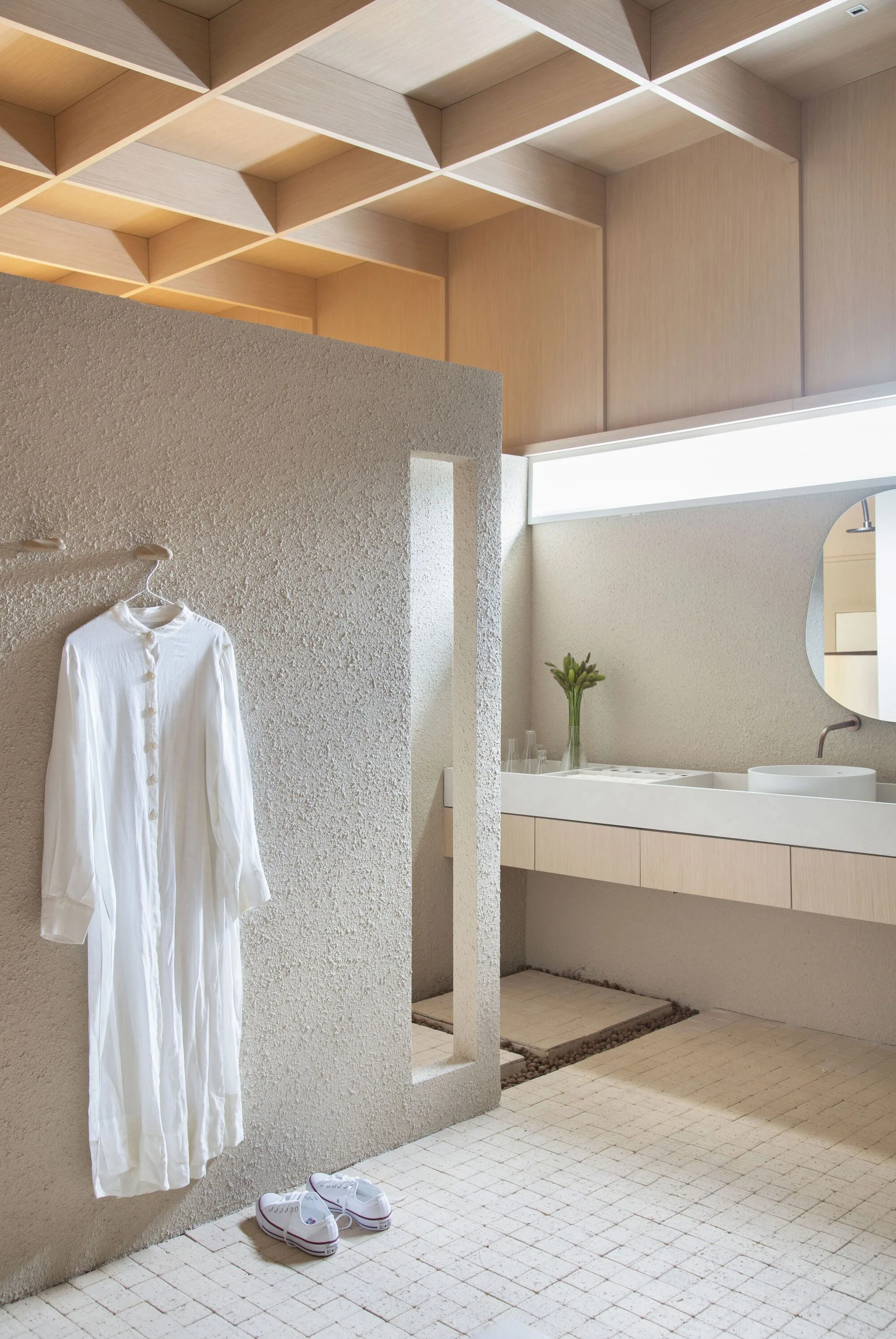 A minimalist bathroom features a rough textured plaster partition wall, a floating vanity with a round mirror, and a light wood grid ceiling.