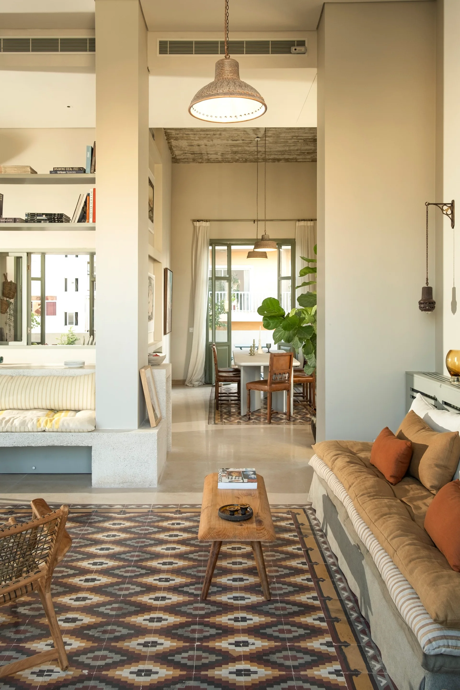 A sunlit living room featuring patterned geometric floor tiles, a tan sofa with rust pillows, a wooden bench coffee table, and built-in plaster shelves.