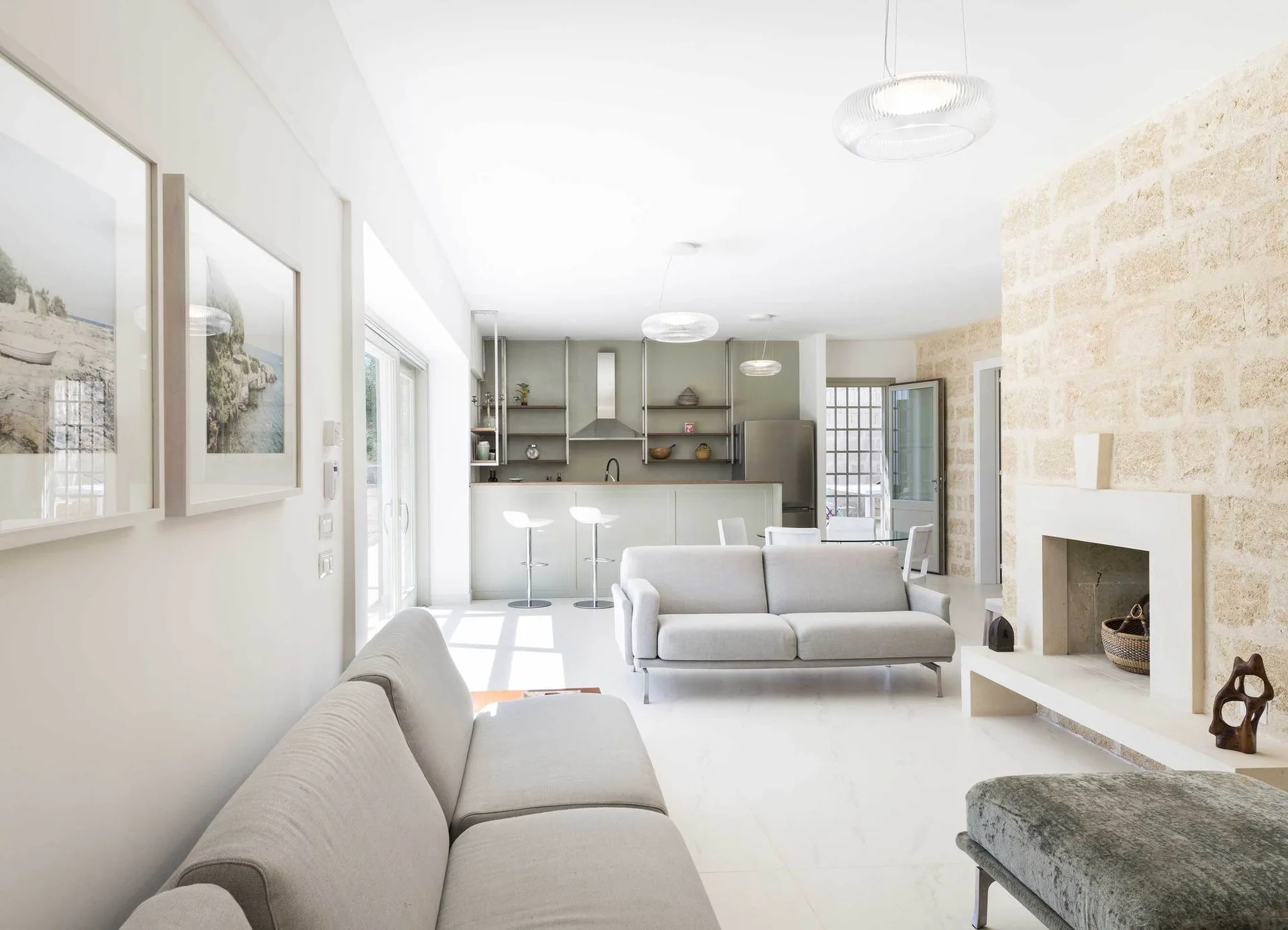 A bright open-concept living room features two light grey sofas, a textured stone fireplace wall, and a pastel green kitchen with bar stools.