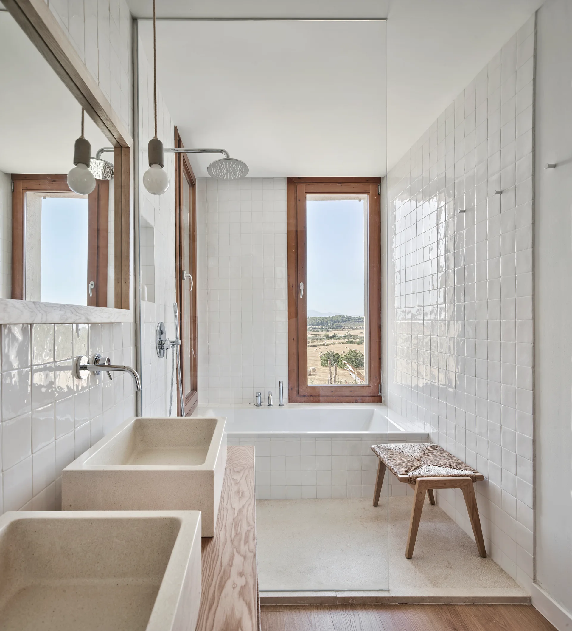 A bright bathroom features white square tiles, a wooden vanity with two stone-like basin sinks, a built-in tub under a wood-framed window, and a woven bench.