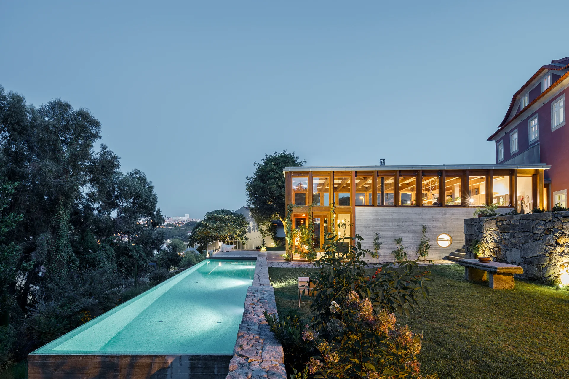 A long rectangular swimming pool glows at dusk next to a lawn and a modern house featuring a double-height glass facade and rustic stone walls.