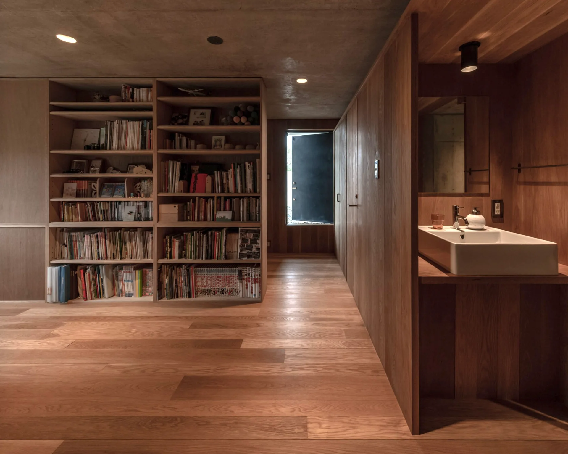 A wood-paneled hallway features a large floor-to-ceiling bookcase, a built-in wooden vanity with a white ceramic sink, and an exposed concrete ceiling.