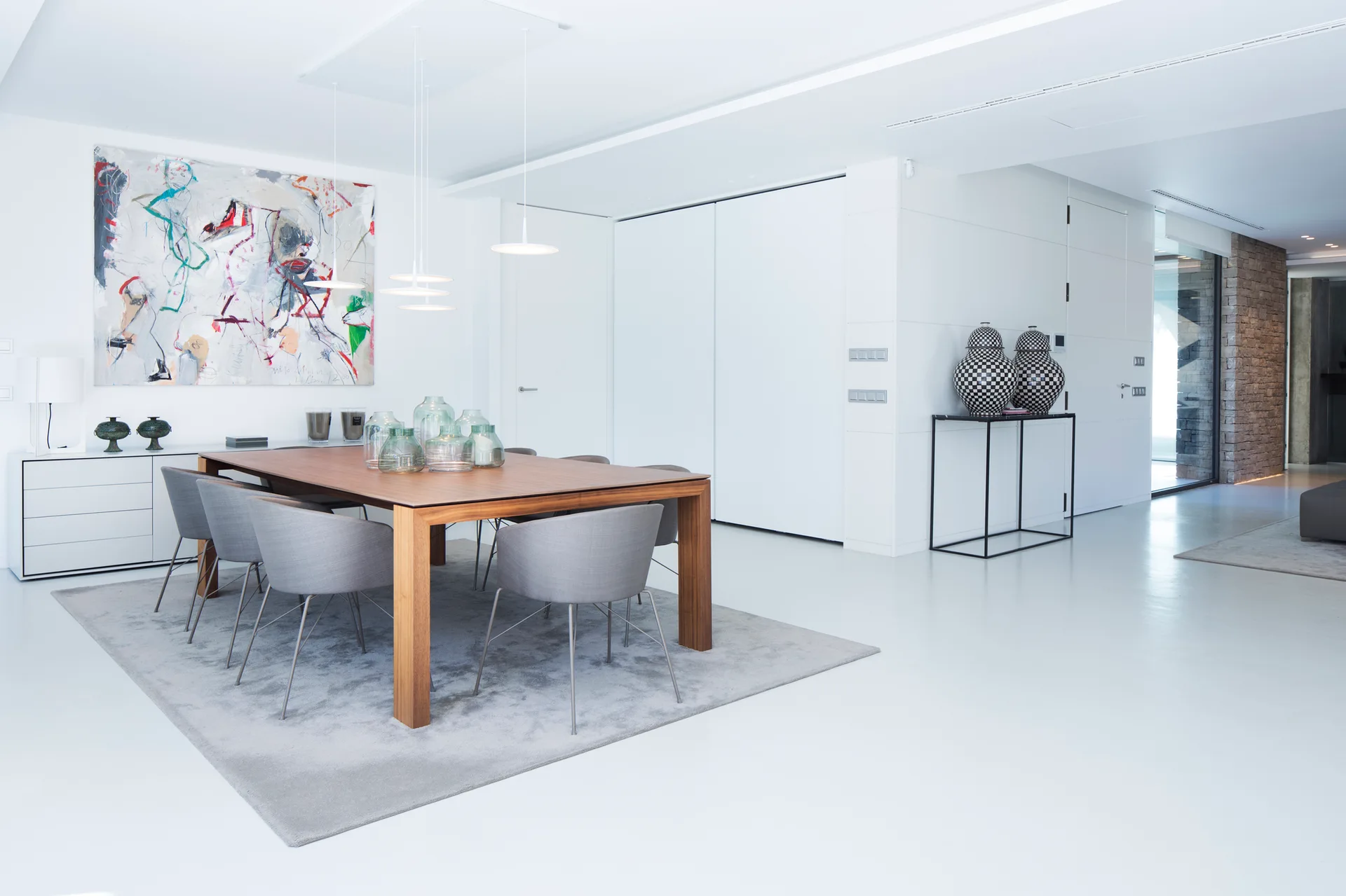 A modern dining room with a rectangular wooden table, grey upholstered chairs on a grey rug, white walls, and a large abstract painting.