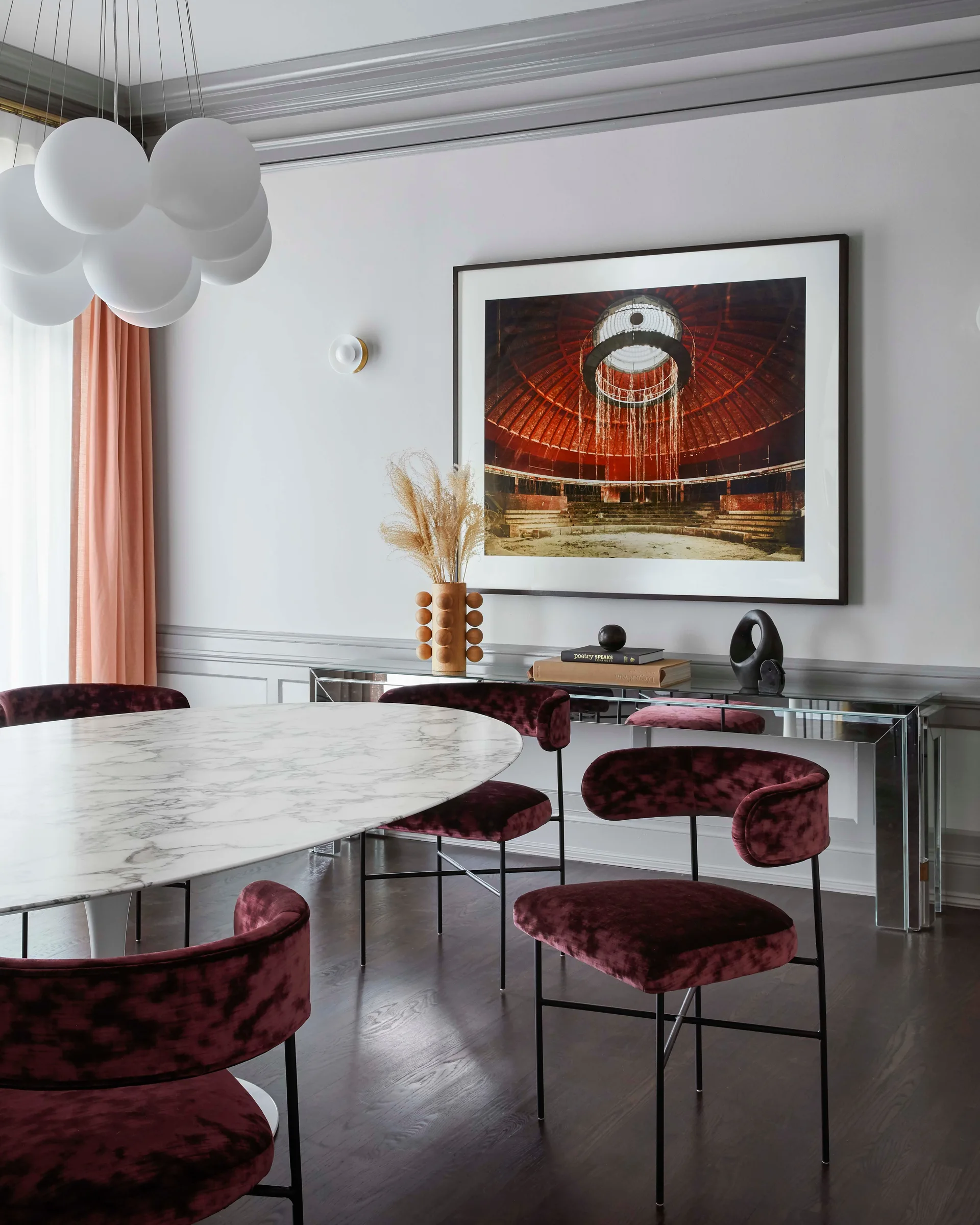 A dining room features a marble-topped oval table surrounded by deep red velvet chairs next to a mirrored console table and a grey paneled wall.
