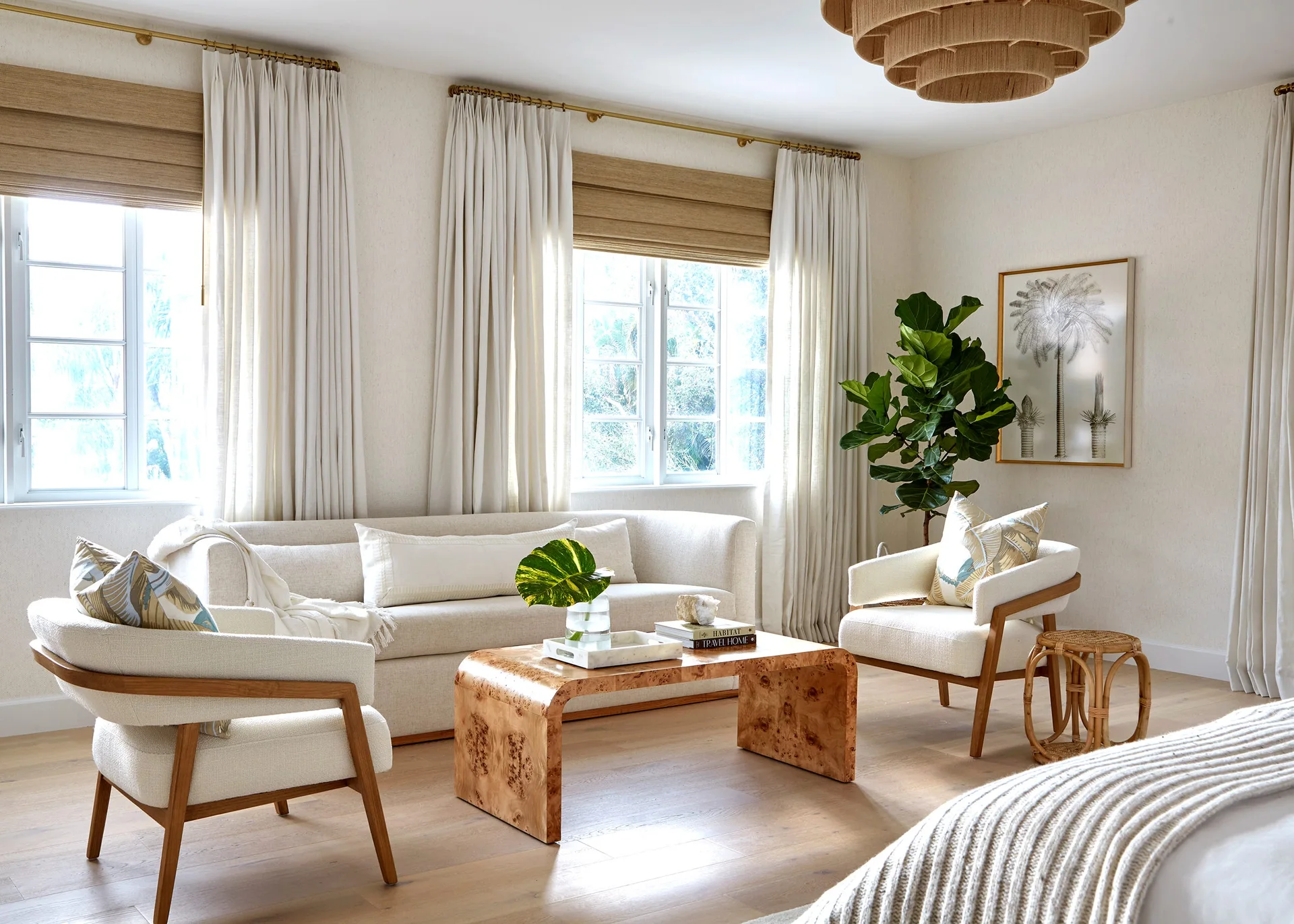 A sunlit bedroom sitting area featuring an off-white sofa, two wooden armchairs, a burl wood waterfall coffee table, and layered window drapes.