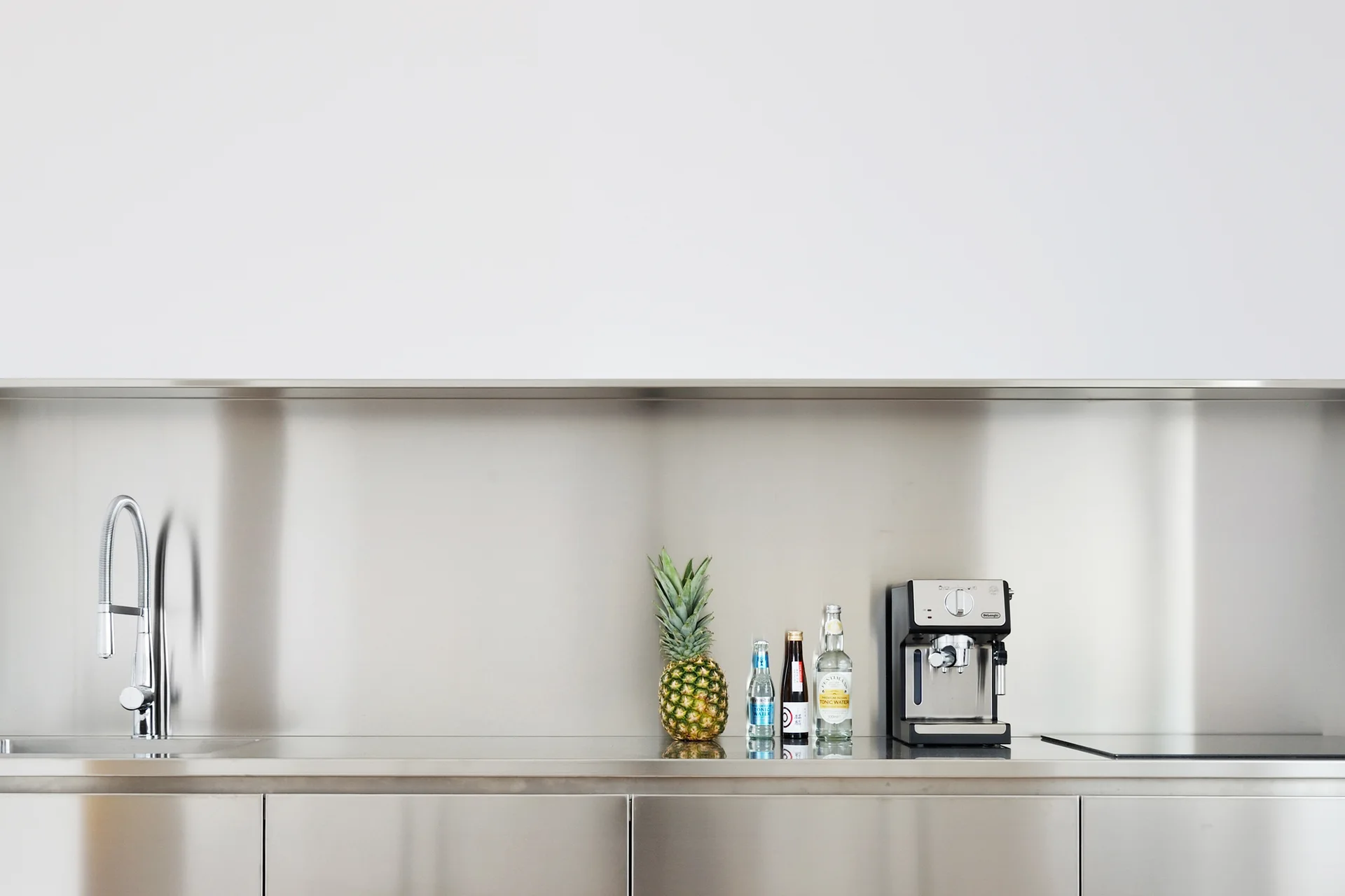 A minimalist kitchen counter with a stainless steel worktop and backsplash featuring a gooseneck faucet, a pineapple, and a silver coffee machine.