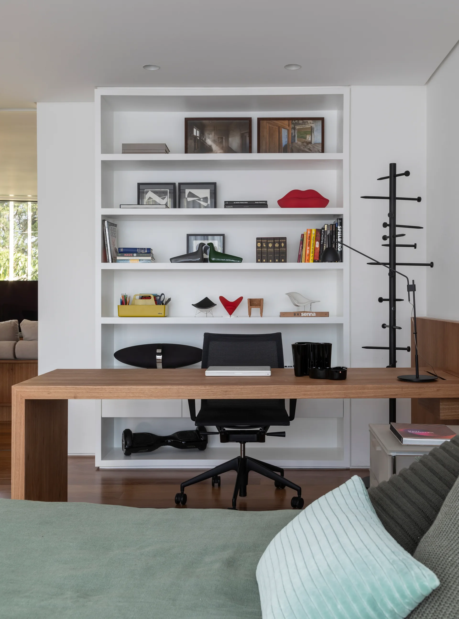 A minimalist home office features a light wood desk, a black mesh chair, and a large white built-in shelving unit filled with books and decor.