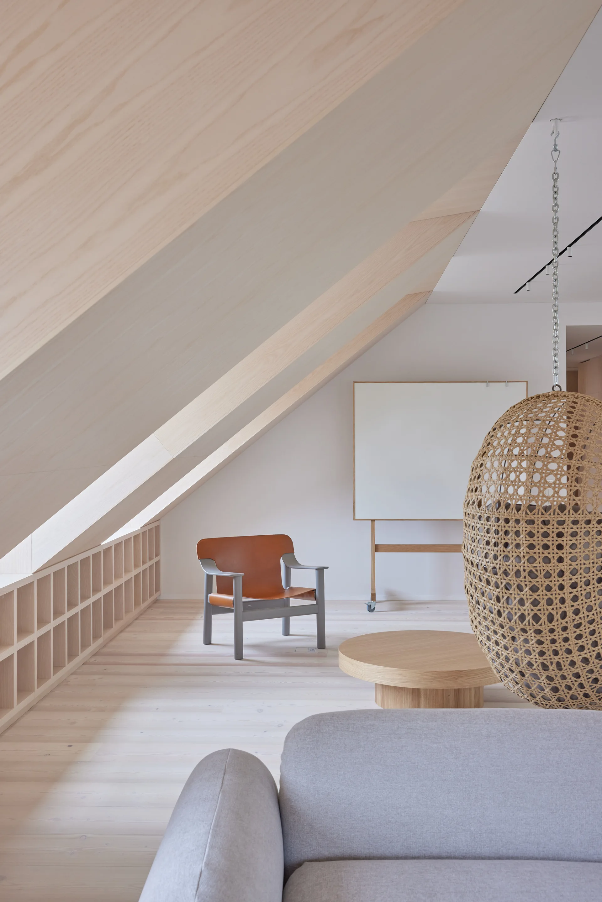 A minimalist attic living room features a slanted wood ceiling, low built-in shelving, an orange leather armchair, and a hanging wicker pod chair.