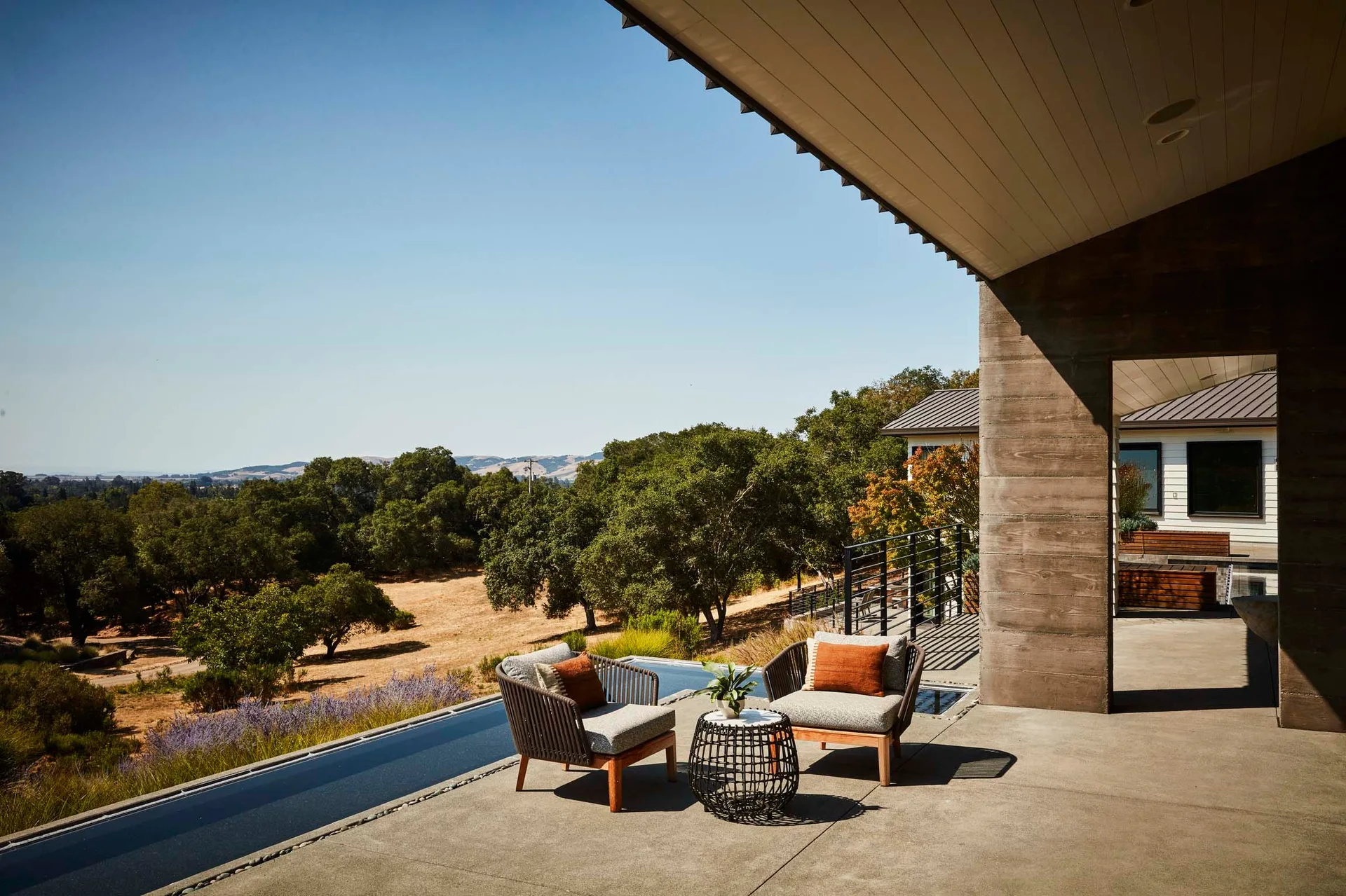 Two grey outdoor lounge chairs with rust pillows sit on a concrete patio next to an infinity pool overlooking a tree-lined valley.