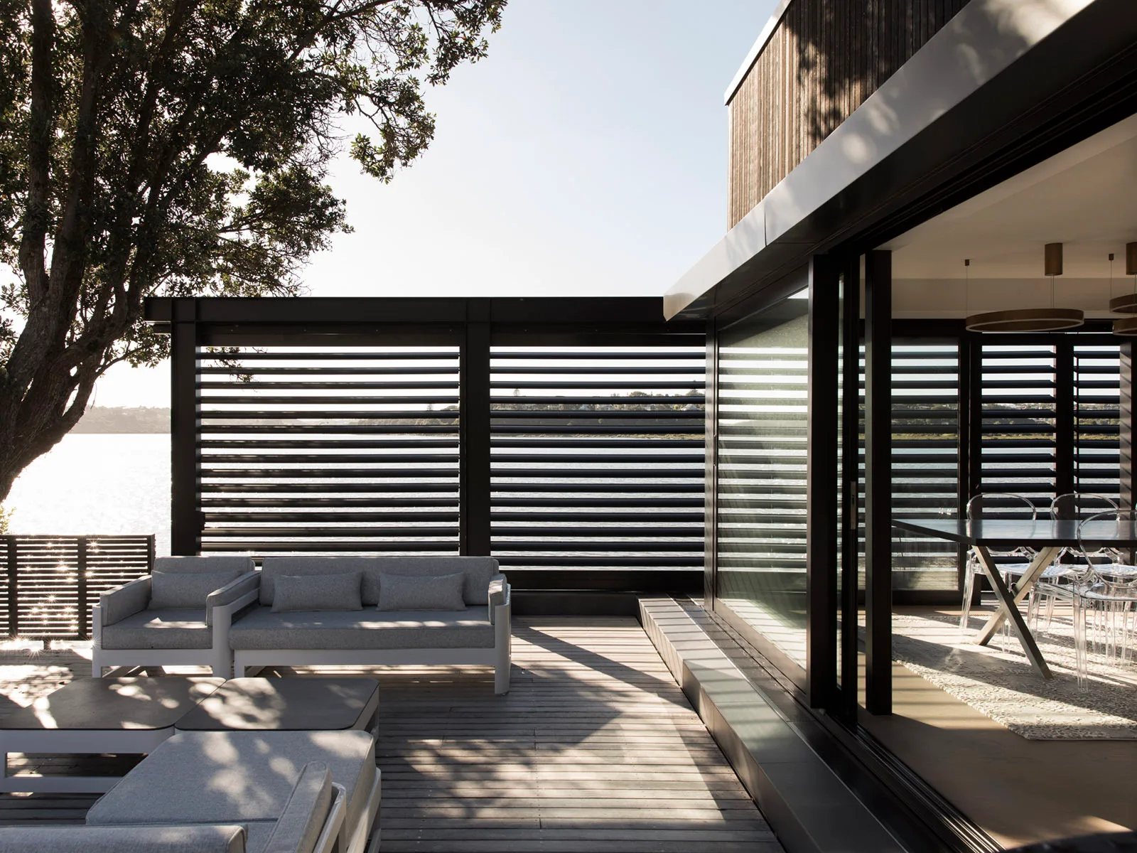 A wooden deck with grey outdoor sofas and black louvered privacy screens next to sliding glass doors that open to an indoor dining space.