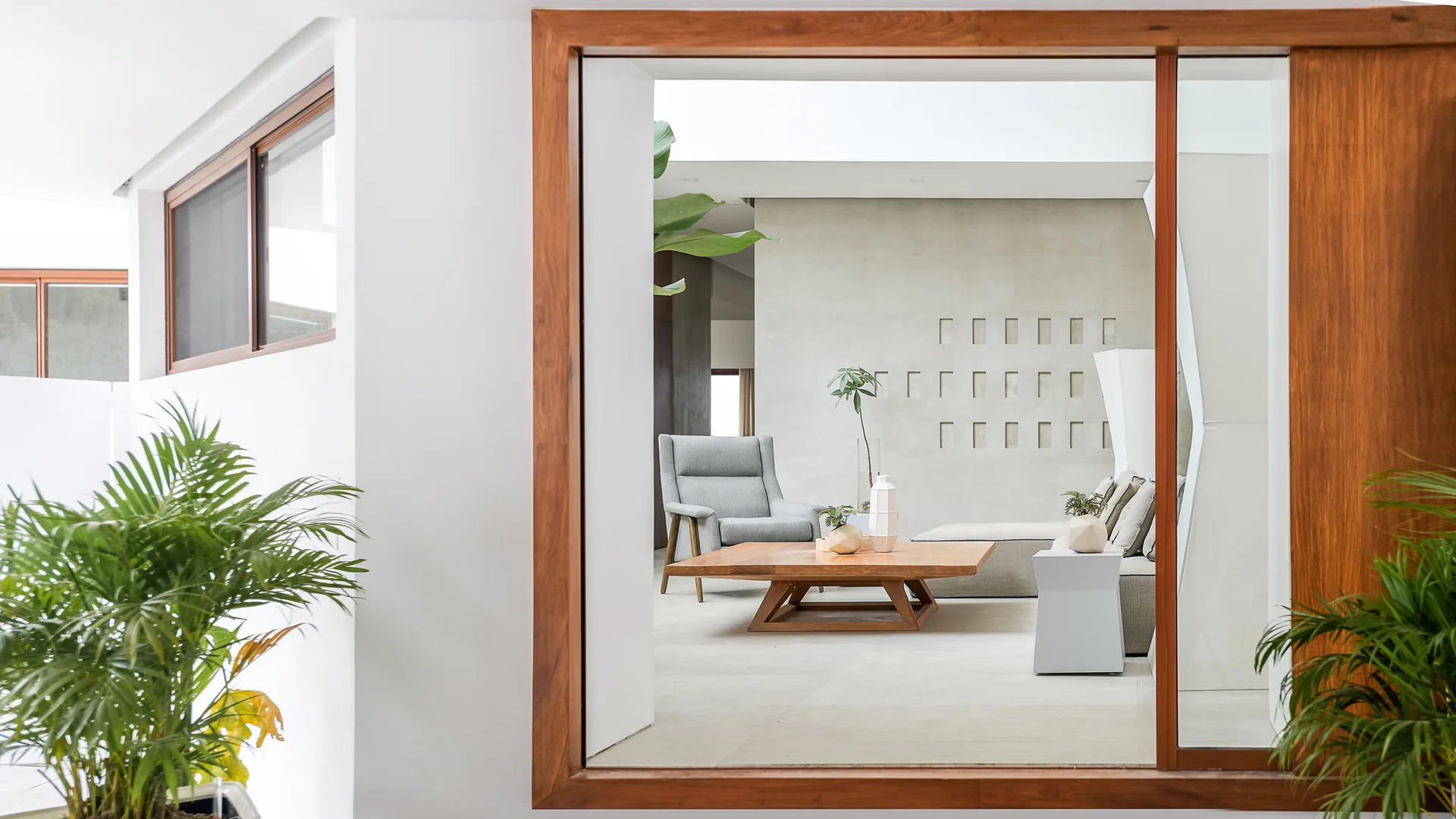 A modern living room with a grey armchair, a wooden coffee table, and a cutout feature wall, viewed through a large wooden window frame.