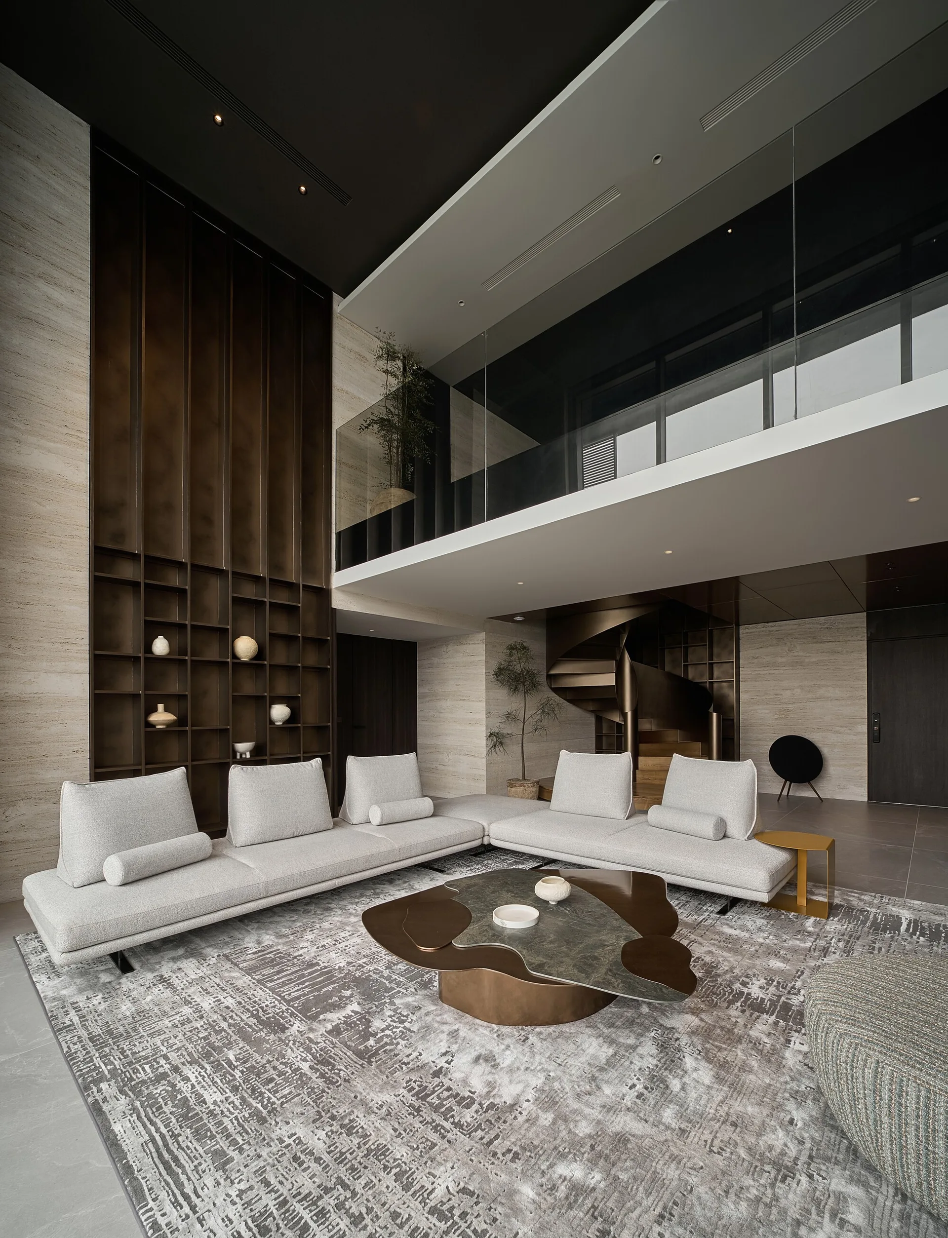 A double-height living room with a grey modular sectional, organic coffee table, tall bronze grid shelving, and a metal spiral staircase.