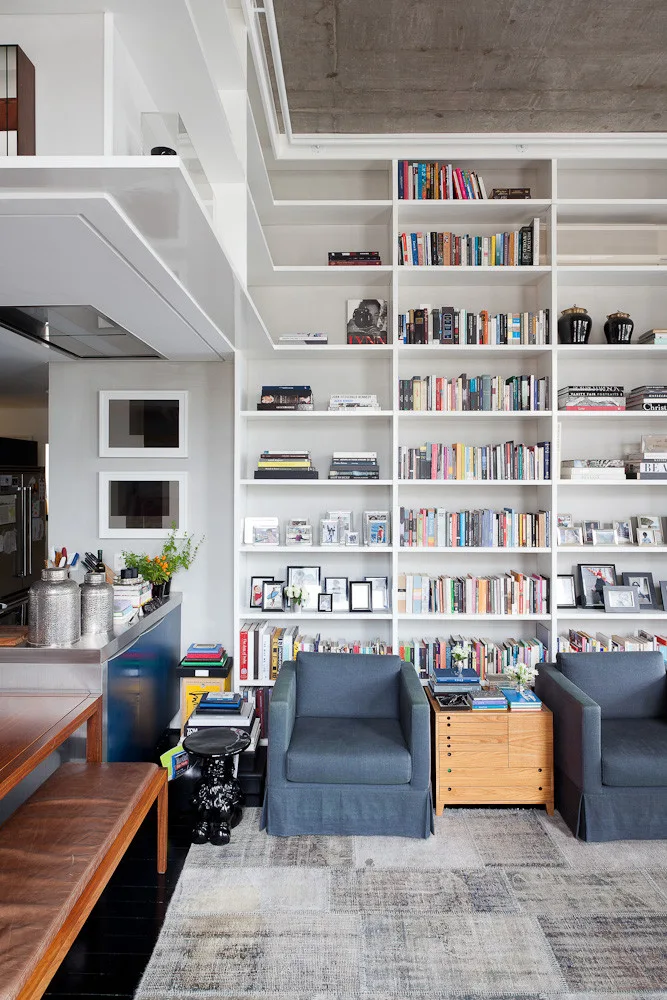 A floor-to-ceiling white bookshelf filled with books stands behind two blue upholstered armchairs on a patchwork rug under a concrete ceiling.